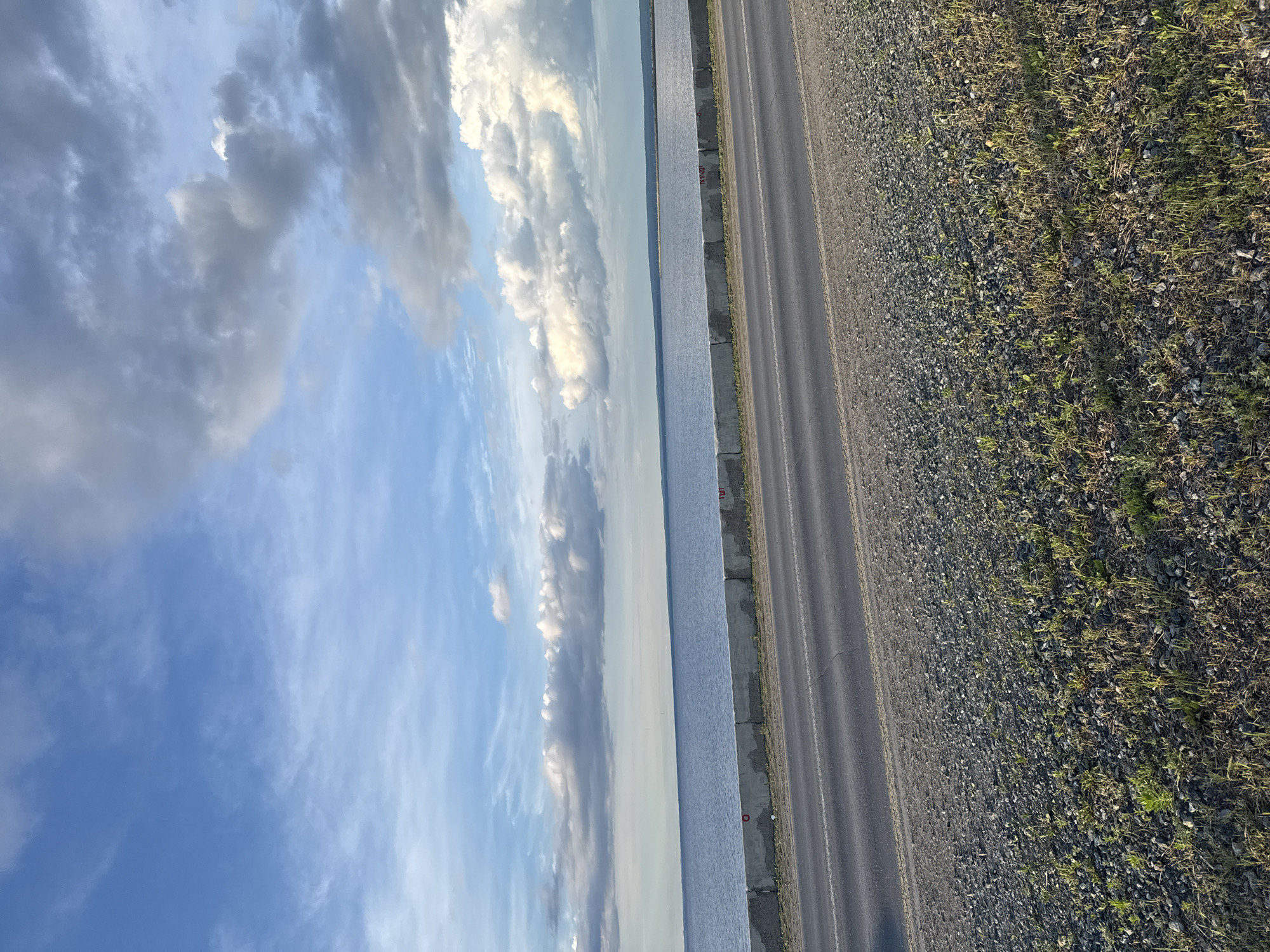 The image shows a view of the Kama River in Chaikovsky, Russia. The photograph appears to be taken from a high vantage point, possibly an embankment or bridge overlooking the river and adjacent infrastructure.
The left side of the frame predominantly features a cloudy blue sky. To the right of the sky is the Kama River. Adjacent to the river, along the right side of the frame, there is a road surface, with a strip of gravel and vegetation alongside it.