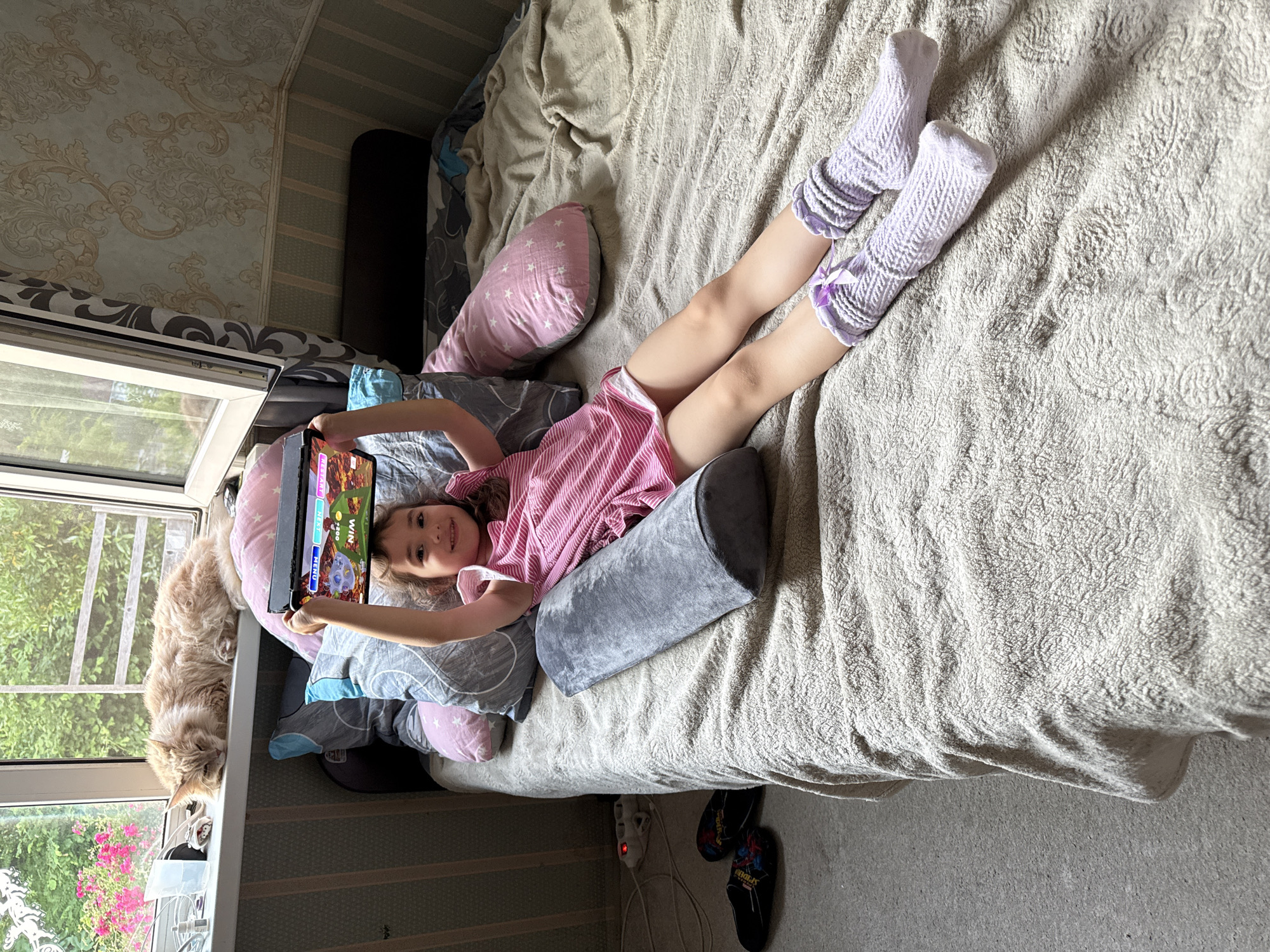 A young girl is lying on a bed, holding a tablet computer. She is wearing a pink striped dress and lilac leg warmers. The bed is covered with a textured beige blanket and several pillows. A large, fluffy ginger cat is lying on a windowsill to the left of the frame, next to a window that looks out onto greenery and some pink flowers.
The tablet screen shows a game with colorful graphics, including cartoon characters and text. Some of the visible text on the screen includes "WIN" and "MENU".
The setting appears to be a bedroom, with patterned wallpaper on the wall behind the bed. The overall atmosphere is relaxed and casual, suggesting a lazy day indoors. The lighting indicates it is daytime. The location context is Kroyanskoye, Russia.
