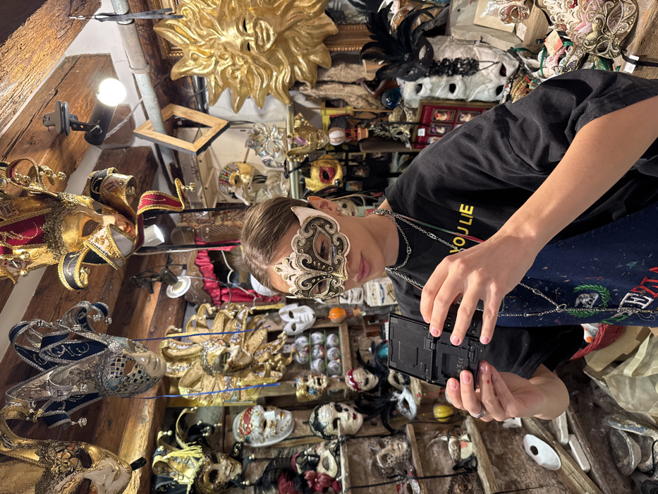The image displays the interior of a mask shop, specifically identified as being in Venice, Italy, due to the distinctive style and abundance of Venetian carnival masks. A young individual with light brown hair, pulled back, is centrally positioned, wearing an ornate white and gold eye mask with dark embellishments. They are dressed in a dark t-shirt with a fragment of yellow text visible and a dark, paint-splattered apron, accessorized with several silver-colored chain necklaces. Both hands, displaying pink-painted fingernails, hold a small, black, open portable electronic device, possibly a gaming console, oriented as if being viewed or operated. The cluttered shop environment is densely packed with a wide variety of Venetian masks, including elaborate jester-style masks, simpler white masks, and a prominent golden sun-face mask, all displayed on wooden shelving units and hanging from rustic wooden ceiling beams. Artificial lighting illuminates the scene.