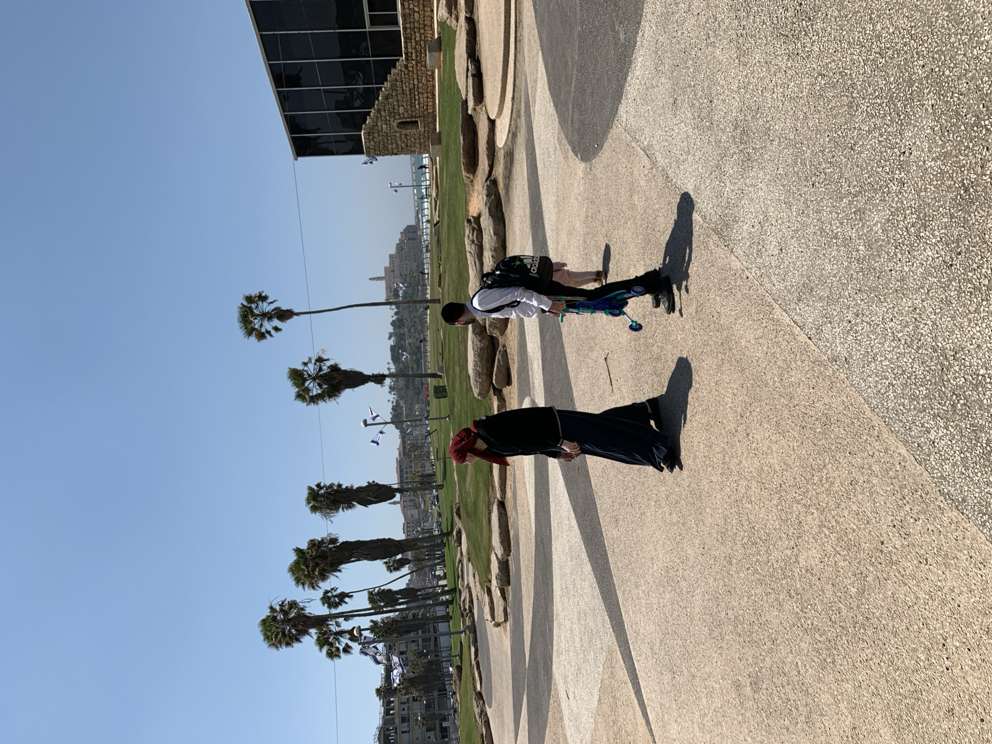 The image shows a sunny outdoor scene in Tel Aviv, Israel, on a paved walkway with palm trees lining a grassy area. In the distance, buildings are visible. Two people are walking on the path. One individual is wearing a white shirt and carrying a black Adidas bag while pushing a blue scooter. The other person is wearing a dark outfit and a red head covering. Israeli flags are hanging from poles along the walkway. A building with glass windows is seen on the upper left.