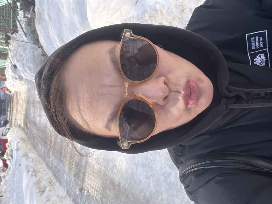 An adult female is depicted in a medium-close-up selfie, standing outdoors in a winter environment in Chaikovsky, Russia. She is wearing a black hooded jacket with a white "Kappa AUTHENTIC SPORTSWEAR SINCE 1967" logo on the right chest area and light-framed, dark-lensed sunglasses. Her lips are puckered in a pouting expression. Her brown hair is visible beneath the hood. The background reveals a partially cleared road or path, covered in a mixture of melting snow, ice, and slush, with tire tracks visible. High banks of dirty snow line both sides of the path. In the middle distance, vehicles, including a red car, green structures, and individuals in orange high-visibility clothing, are discernible. Stairs leading up to a structure are also visible on the right. The scene is illuminated by bright sunlight, casting reflections on the wet surfaces.