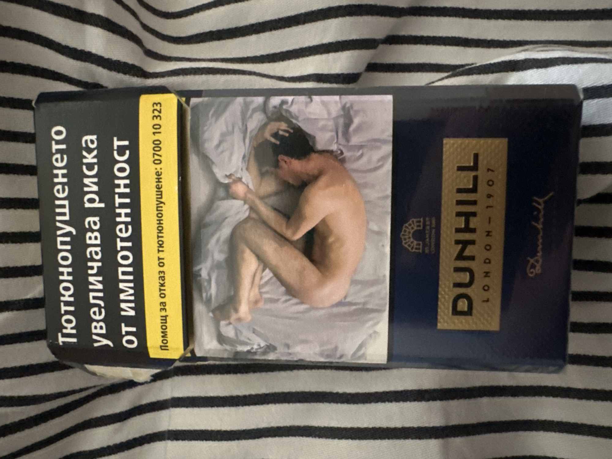 The image shows a pack of Dunhill cigarettes placed on a striped fabric. The top of the pack displays a warning in Bulgarian text about the risks of smoking, specifically mentioning increased risk of impotence. Below the warning, there's a yellow band with a phone number for smoking cessation assistance. The middle section of the pack features a photograph of two people in bed, appearing intimate. The bottom part of the pack has the Dunhill logo and "LONDON 1907" printed in gold on a dark blue background. The background fabric consists of alternating black and white stripes. The overall setting is indoors, with the pack resting on a piece of fabric.