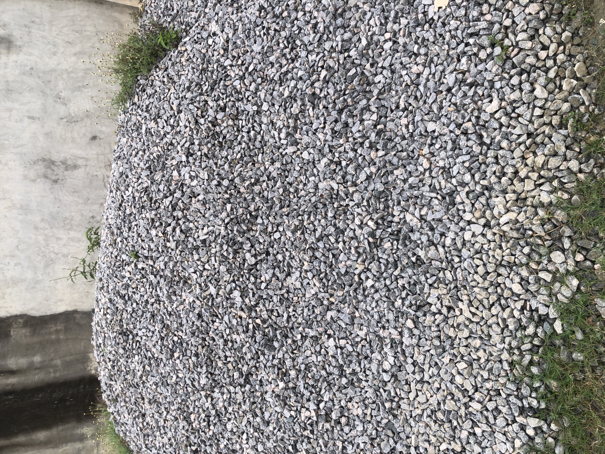 A large pile of crushed stone, likely for construction, sits against a rough, weathered concrete wall. The stone is a mix of gray, black, and white fragments, forming a mound that fills most of the frame. Sparse patches of green grass and a few small wildflowers grow along the bottom edge of the pile and near the wall. The concrete wall appears to be a section of a building or a retaining wall, with a darker, damp-looking area on the left side. The scene suggests an outdoor location in Ado, Nigeria, possibly a construction site or a material storage area. There are no people visible in the image.