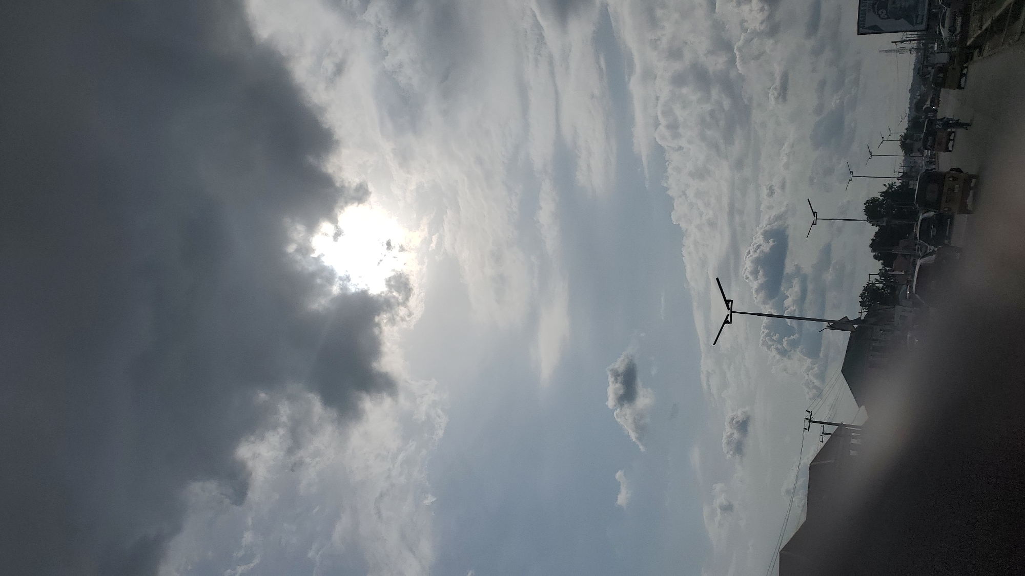 The image depicts a partially cloudy sky over a street scene in Jalingo, Nigeria. The sun is visible, partially obscured by dark, heavy clouds on the left side of the frame, suggesting either early morning or late afternoon. The clouds are a mix of dark, ominous ones and lighter, more diffused ones, indicating a dynamic weather pattern.
The scene on the ground shows a street with several vehicles, including what appear to be auto-rickshaws (a common mode of transport in Nigeria) and other cars. Streetlights with multiple fixtures are visible, lining the road. Trees and buildings can be seen in the background, with some of the buildings appearing to be simple structures. The overall impression is of a street in an urban or semi-urban area during the daytime.
There is some text visible in the upper right corner of the image, on what looks like a billboard or sign, but it is too blurry to be deciphered. The image is oriented vertically, as if it was taken while holding a phone in portrait mode. No people are clearly visible or identifiable in the image. The weather appears to be transitioning, with the heavy clouds suggesting potential rain, but the presence of the sun indicates it is not currently raining heavily.