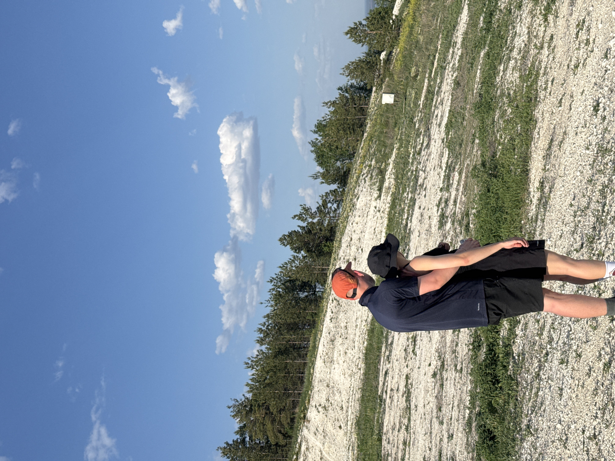 Two people are standing on a gravel path alongside a hill covered in trees, under a blue sky with scattered clouds. One person wears an orange baseball cap, a dark blue shirt, and black shorts, while the other wears a black baseball cap and is embracing the first person. The location is likely near a chalk mountain, a geological feature common in the Khvalynsk region of Russia.