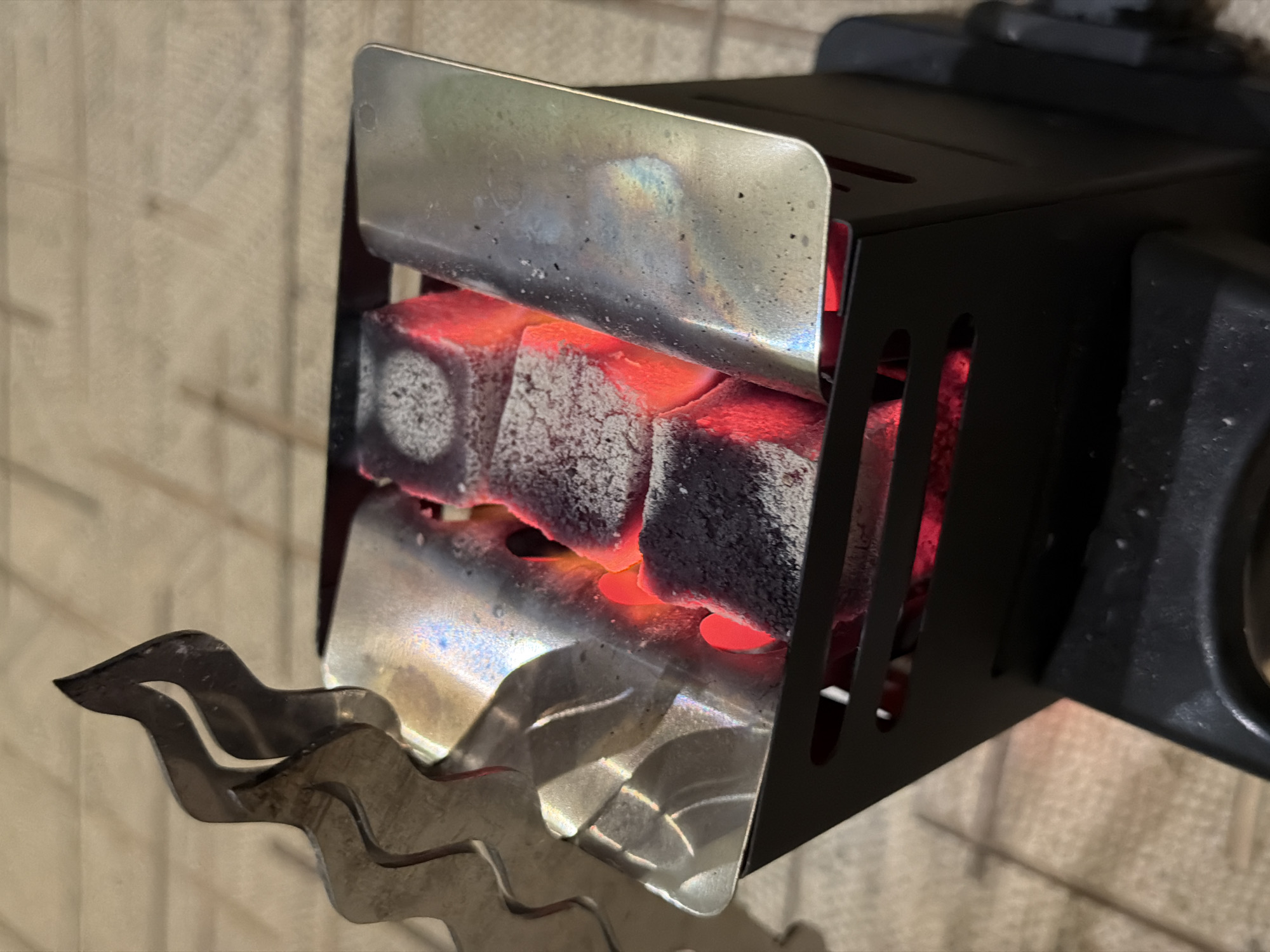 A close-up shot reveals a black metal charcoal heater with three glowing-red charcoal cubes inside. The heater has a front shield of shiny metal with rainbow-like heat discoloration and a decorative wavy edge. The heater is positioned against a wall with a wood-like pattern. The image does not show any people, actions, or interactions. The location is not discernible in the image.