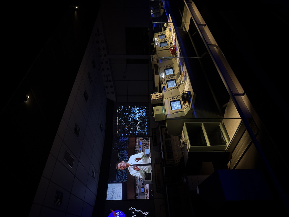The image depicts a dimly lit, simulated mission control center, specifically located within an exhibition in Canela, Brazil. The scene features a large projection screen on the rear wall displaying a male presenter in a white collared shirt and dark tie, gesturing with both hands, flanked by graphical data, including a NASA logo and complex charts. In the midground and foreground, a series of vintage-style control consoles, light gray in color, are arranged. Each console is equipped with multiple square CRT monitors displaying blue graphical information. Objects on the consoles include a black rotary telephone, a red push-button telephone, and stacks of papers. A clear glass barrier separates the viewer's area from the control consoles. Overhead lighting fixtures are visible on the dark ceiling, casting focused illumination onto the consoles and creating reflections on the glass barrier. The overall environment suggests a museum exhibit designed to recreate a historical space operations setting. There are no live individuals present within the frame beyond the projected image.