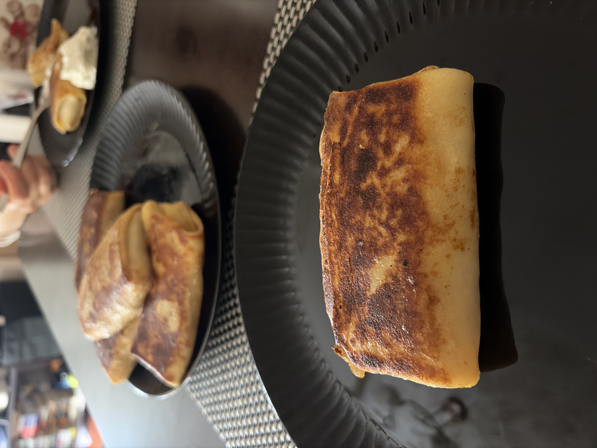 The image shows three black plates on a surface. The surface is covered with a checkered patterned mat. A single rolled crepe is on a plate to the right. A plate with three crepes is situated on the left. There is a plate containing rolled crepes with a dollop of white cream on the upper left. The location, Budva, Montenegro is not discernible from the image.