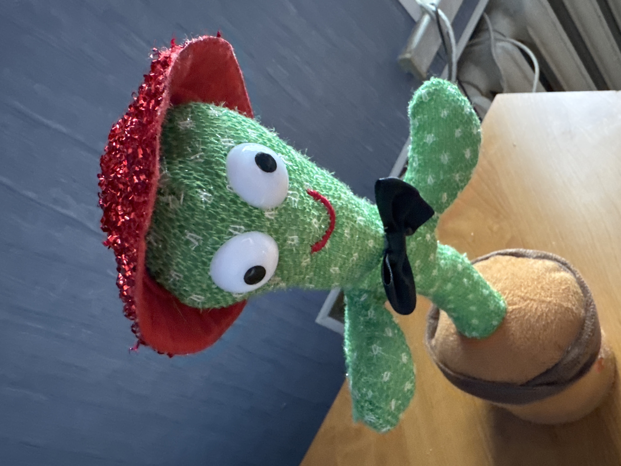 A close-up, low-angle shot shows a plush toy cactus with large, googly eyes and a red, sequined hat. The cactus has green fabric with white dots and is wearing a black bow tie. Its arms are outstretched, and it is planted in a brown, fuzzy pot. The cactus is positioned on a wooden surface, with a blue, textured wall in the background. Electrical wires are visible to the right of the cactus.