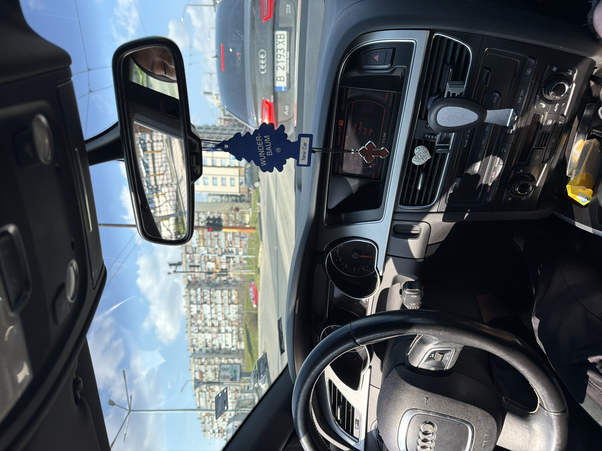 The image depicts a first-person view from the interior of a stationary Audi vehicle, looking forward through the windshield at a city street and intersection.
Inside the vehicle, the black Audi steering wheel with its four rings logo is prominent on the left. The instrument cluster shows the speedometer at 0 km/h and the tachometer at idle. The infotainment screen displays "Radio FM" with options such as "News," "Culture," "Pop," and the time "15:34." A blue, tree-shaped "Wunder-Baum New Car" air freshener and a silver-and-red cross charm hang from the rearview mirror. The rearview mirror itself reflects a partial view of a person's face, likely an occupant. A sparkling heart-shaped ornament and a mobile phone holder are attached to the central air vents. Below the climate control panel, a yellow Orbit chewing gum package and a dark rectangular object are visible in a storage compartment. The driver's legs, clad in dark trousers, are visible below the steering wheel.
Outside the vehicle, the road ahead includes a white stop line. A dark grey Audi A8 sedan is stopped directly in front. Its Bulgarian license plate, "B 2193 XB," is clearly visible, with "B" indicating Varna. The car's red taillights are illuminated. A traffic light to the right is red. In the background, large, multi-story residential buildings with numerous balconies, characteristic of urban Bulgarian architecture, line the horizon under a clear blue sky with scattered white clouds. Streetlights, utility poles, a green grassy median, a distant red car, and a billboard displaying the number "8" are also present.
The scene is an urban intersection in Varna, Bulgaria, characterized by the specific architecture and the local license plate.