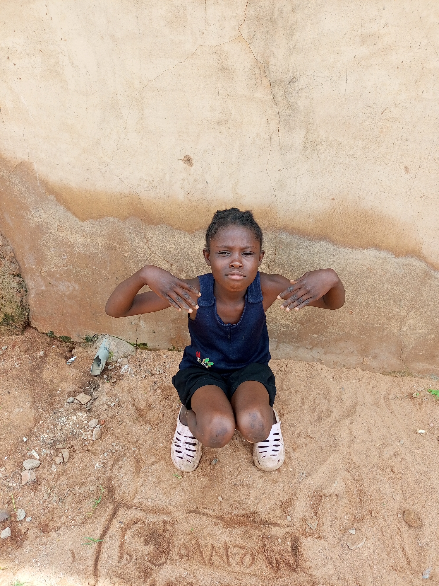 In Tempure, Nigeria, a young girl kneels on sandy ground in front of a weathered, beige-colored wall. The wall shows signs of age, with cracks and patches of discoloration, particularly a brown stain that streaks diagonally across it. At the base of the wall, a discarded white pipe protrudes from the ground, surrounded by dirt and small stones. The girl, dressed in a navy blue tank top with a small cartoon character on it and black shorts, has her hands raised, palms facing outward, in a gesture of defense or perhaps a playful pose. Her knees are bent, and she wears light-colored, open-toed sandals. The sandy ground beneath her is uneven, with some faint markings or footprints visible. The overall scene suggests an outdoor environment in a rural or semi-urban setting.