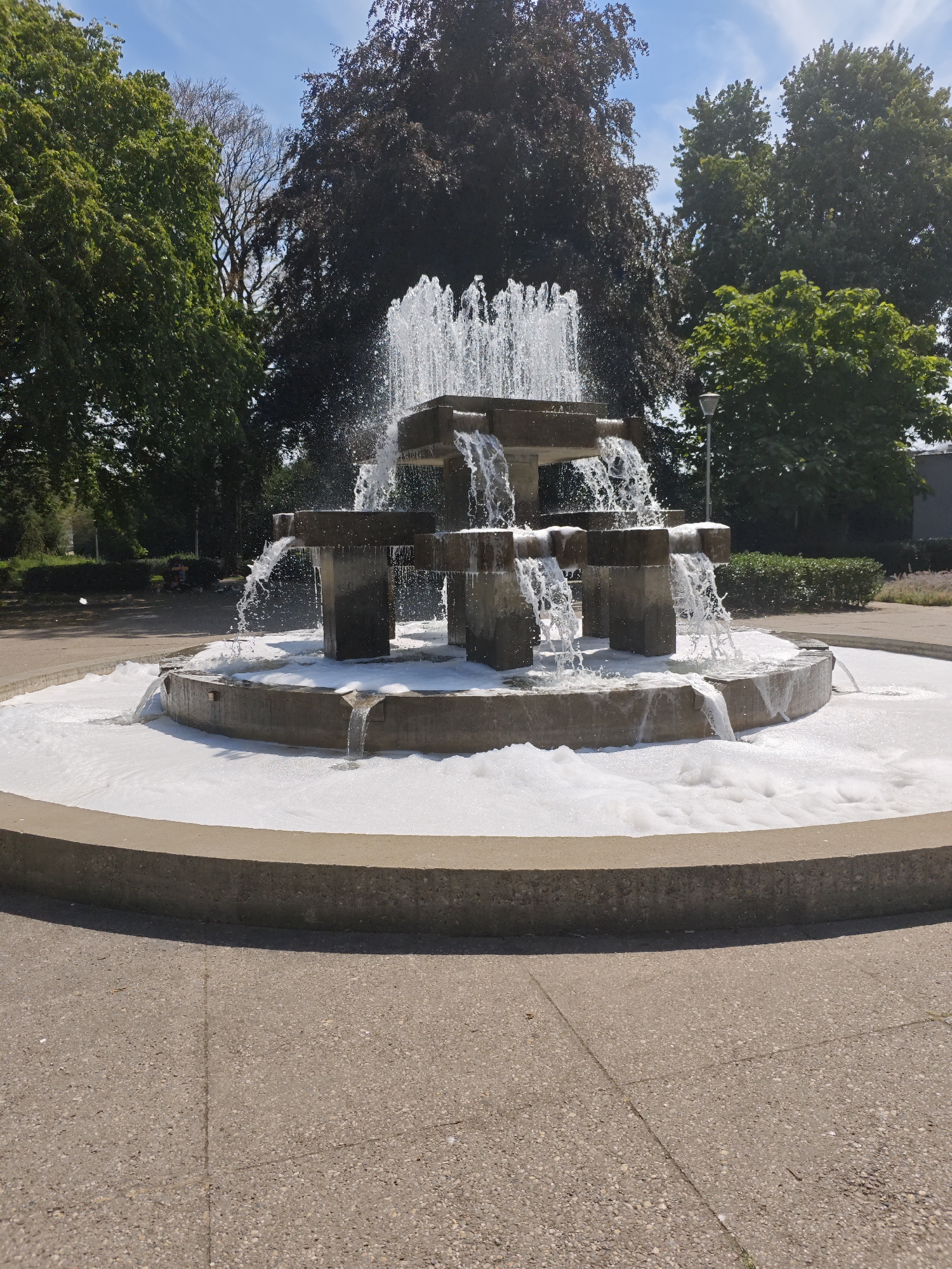 A tiered concrete fountain with frothy white water cascading down its levels is centrally located in a park. The fountain is surrounded by a circular concrete rim, which is itself on a paved pathway. Green trees with dark foliage and some with lighter green leaves form a backdrop, with a clear blue sky above. To the right of the fountain, a modern lamppost is visible behind some foliage. This scene is located in the Burgemeester van Baarspark in Tilburg, Netherlands. The image is a static, medium shot capturing the fountain from a slightly elevated perspective. There are no people or animals visible in the frame.