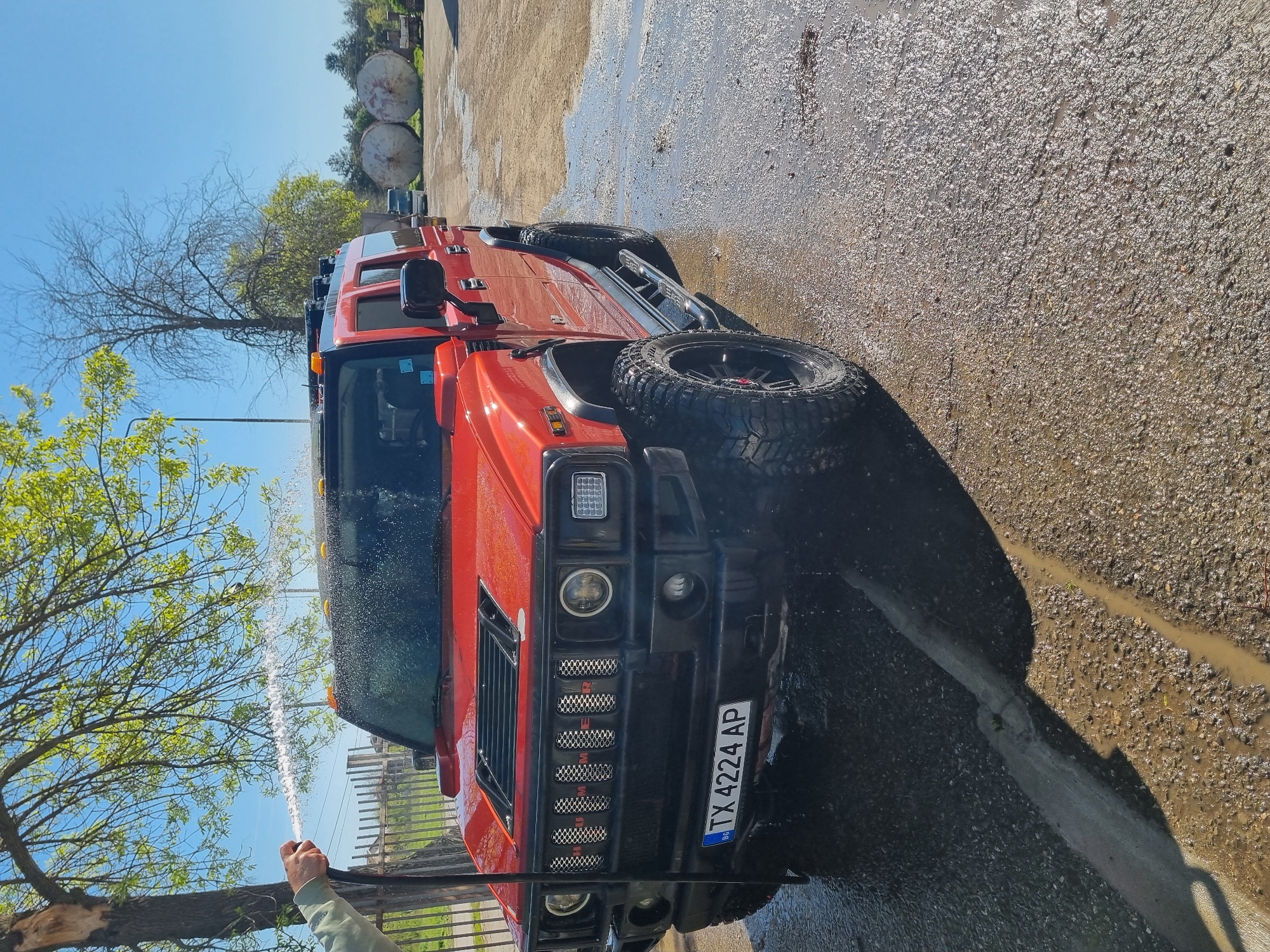 A bright red Hummer H2 SUV is being hand-washed outdoors on a clear, sunny day in Ovcharovo, Bulgaria. The vehicle, positioned prominently in the frame, features large, rugged off-road tires with black rims and a distinctive front grille displaying the word "HUMMER" in individual letters. Square headlights and turn signals are visible on the front. A person's arm, clad in a light-colored sleeve, is visible on the left, holding a hose from which a stream of water is actively spraying onto the vehicle's hood and windshield.
The ground beneath and around the Hummer is a mix of gravel and dirt, appearing extensively wet and muddy with puddles and reflections from the bright sunlight, indicating the ongoing washing activity. A concrete curb or barrier runs along the right side of the wet ground.
In the background, against a clear blue sky, are trees with both bare branches and fresh green leaves, suggesting spring or early summer. Further back and to the right, two large, cylindrical industrial tanks or drums are visible, hinting at an industrial or semi-rural setting. The vehicle's license plate, "BG TX 4224 AP," is clearly legible on the front bumper, confirming its Bulgarian registration. The strong sunlight and clear skies suggest it is daytime, likely morning or early afternoon.