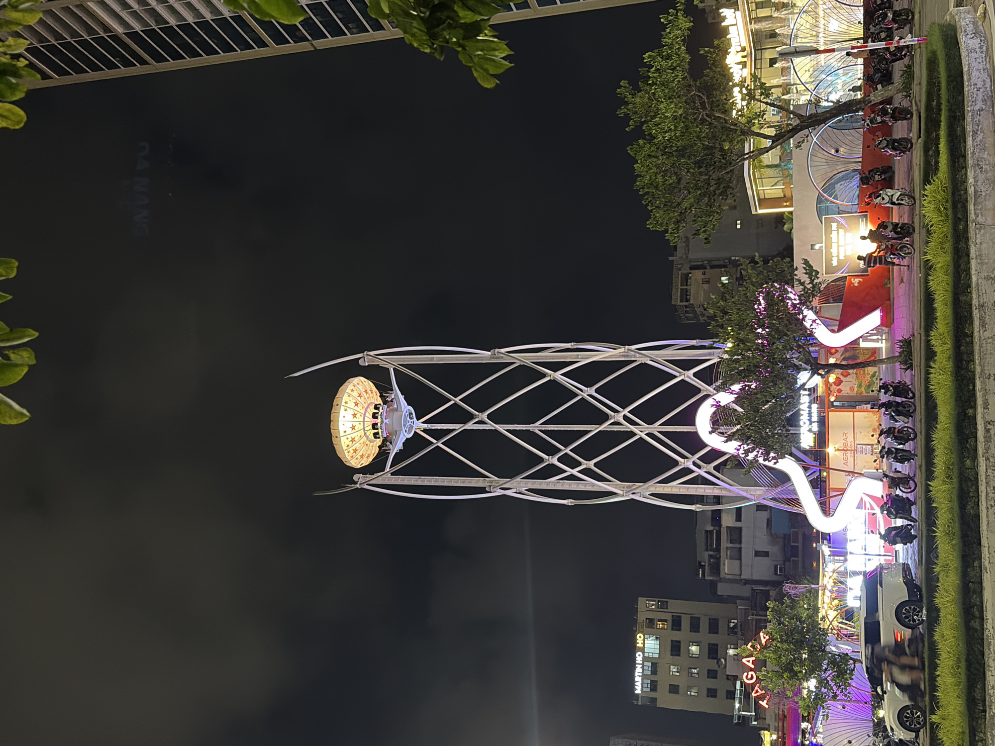 The image depicts an outdoor urban scene at night, likely in Da Nang, Vietnam, as indicated by text on a building that reads "DA NANG". A prominent feature is a tall, white, lattice-like tower structure with a decorative, illuminated orb at its apex, resembling a hot air balloon with star patterns. In the foreground, a white SUV is parked, with several motorcycles also present. The surrounding environment includes buildings with illuminated signs, trees with some glowing purple lights, and decorative arches with white and yellow lights. Several people are visible, some standing near motorcycles and others appearing to be within buildings or the illuminated structures. The overall atmosphere suggests a lively, possibly festive, nighttime environment in a city.