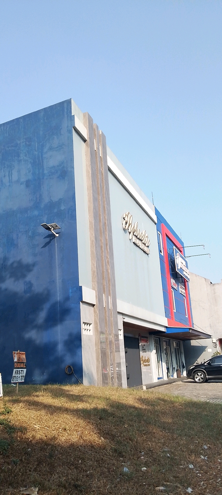 The image captures a building facade with a prominent blue section on the left, transitioning to a lighter blue and gray section adorned with vertical decorative elements. The building features signage with stylized gold lettering that reads "Yupiter". To the right, a section of the building is painted red and blue, with a sign displaying white text on a blue background. Below the building, a grassy area with dry grass is visible. A black car is parked to the right of the building. In the foreground, on the blue wall of the building, there are signs, one of which indicates "DIJUAL" (for sale). The sky is clear and blue. The overall scene depicts a commercial building in an outdoor environment.
