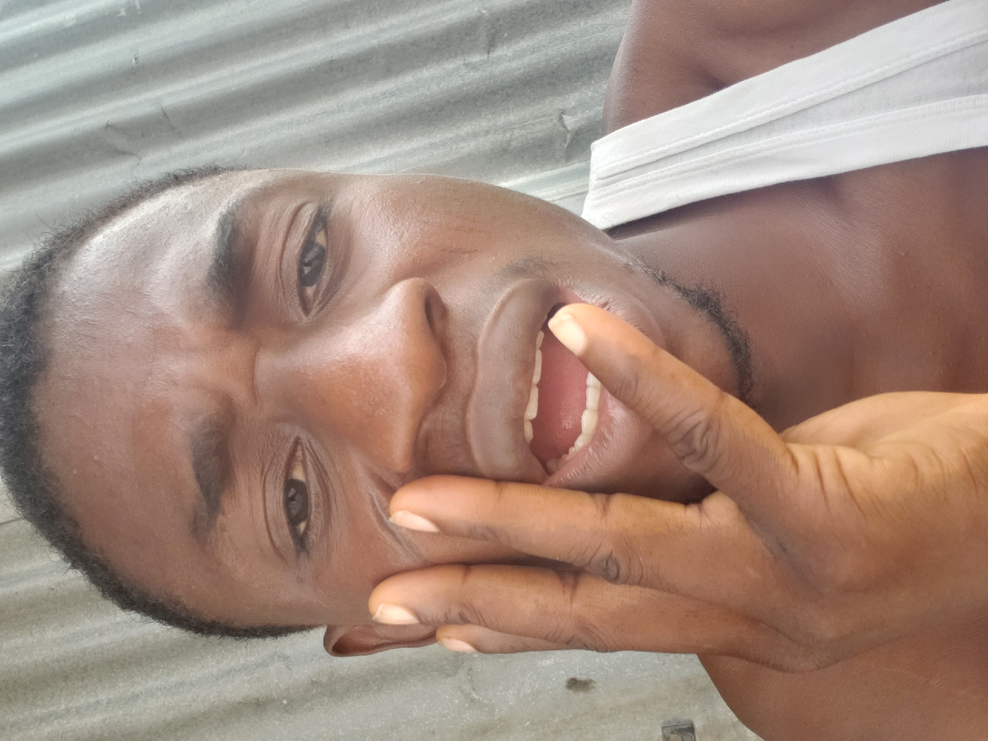 A close-up, tilted shot features a dark-skinned man with short, dark hair and brown eyes. He is wearing a white tank top. His hand partially covers his face, and his mouth is open. The background consists of corrugated metal. The image appears to be taken outdoors in an unknown location, though it is hypothesized to be Adankolo, Nigeria.
