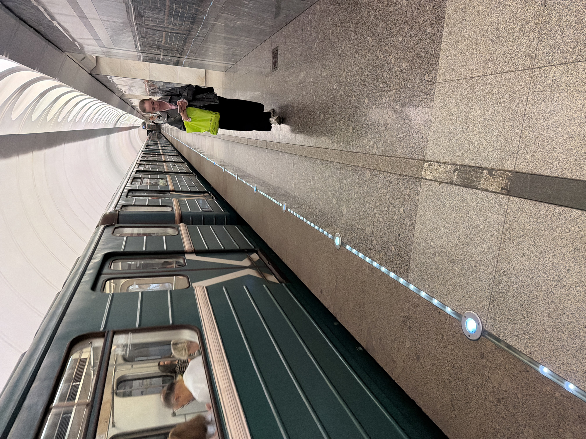 A woman is walking on a subway platform in Moscow, Russia, holding a bright green shopping bag and a smartphone. The platform is lined with a train and illuminated by strip lighting and circular blue lights embedded in the floor. The subway cars are dark green with white accents. The station's architecture features a curved, white ceiling with a pattern of overlapping, translucent panels and marbled walls. Several passengers are visible inside the train cars, seated and looking out the windows. The floor is a speckled gray granite.