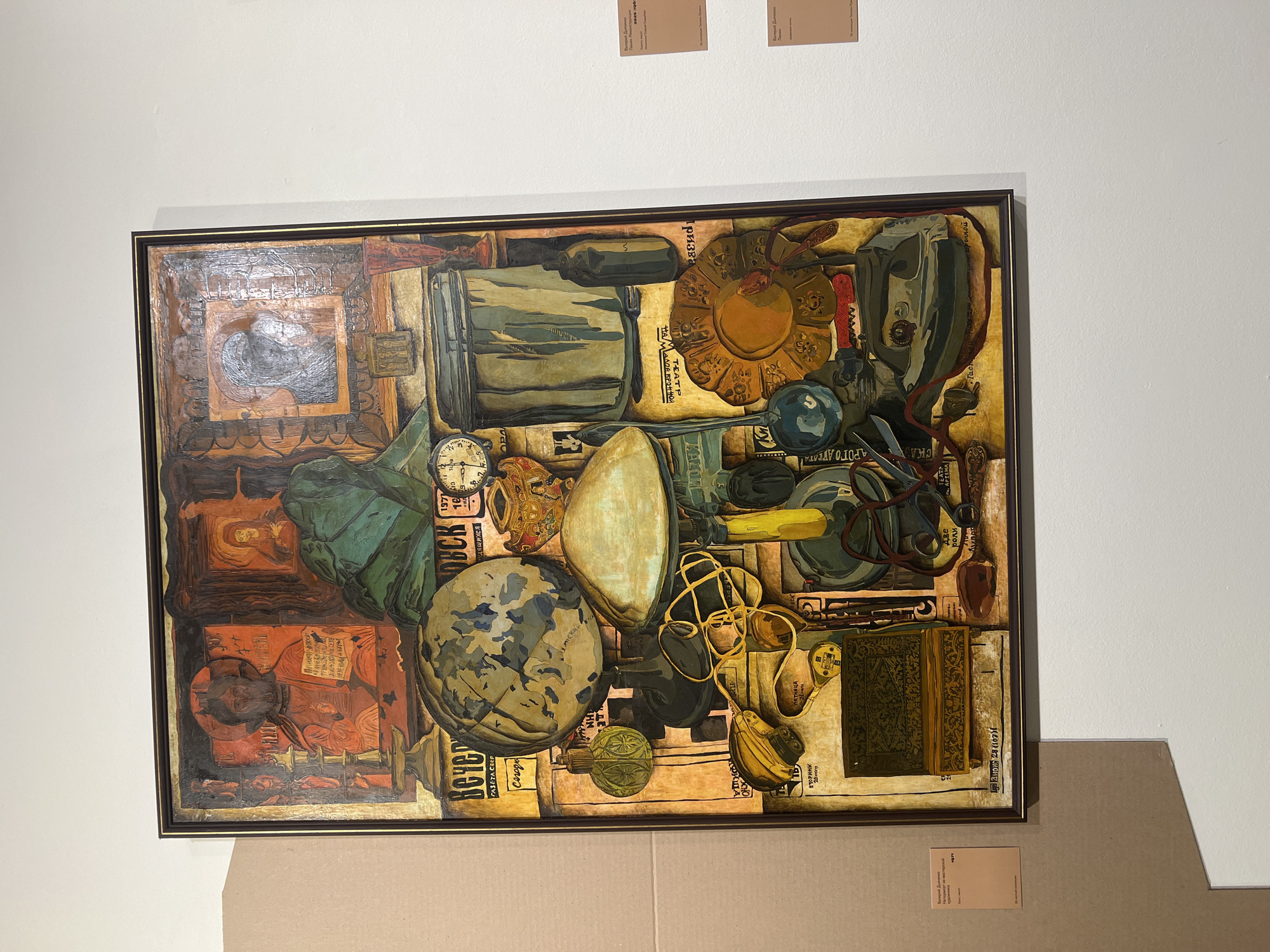 The image shows a framed painting displayed on a white wall. The painting is an oil on canvas, depicting a still life composition with religious icons at the top and a collection of everyday objects below. These objects include a globe, a lamp, an iron, scissors, a clock, and various other items, arranged in a dense and somewhat chaotic manner. The style is figurative with elements of surrealism.
To the left of the painting, a large piece of beige cardboard is leaning against the wall, partially obscuring the edge of the artwork. To the right of the painting, three small informational cards are attached to the wall, with text in Russian. One card, positioned above the painting, is slightly blurred. The card directly to the right of the painting has the title "Вечерний Дмитренко. Попытка. Реконструкция", the text "Холст, масло", and the year "2023". The card below it has the title "Вечерний Дмитренко. Попытка.", and the text "Цифровая копия". A third card, further to the right and partially out of frame, has the title "Вечерний Дмитренко. Попытка. Реконструкция", the text "Холст, масло", and the year "2023". A fourth card, visible at the bottom right, also contains Russian text.
The informational cards suggest that this artwork is likely on display at a museum or gallery in Yekaterinburg, Russia. The title "Вечерний Дмитренко. Попытка. Реконструкция" suggests a connection to an artist named Vecheri Dmitrenko, and potentially a reinterpretation or reconstruction of a past work. The presence of multiple cards with similar titles and dates indicates an exhibition focused on this artist or theme. The environment appears to be an indoor exhibition space.