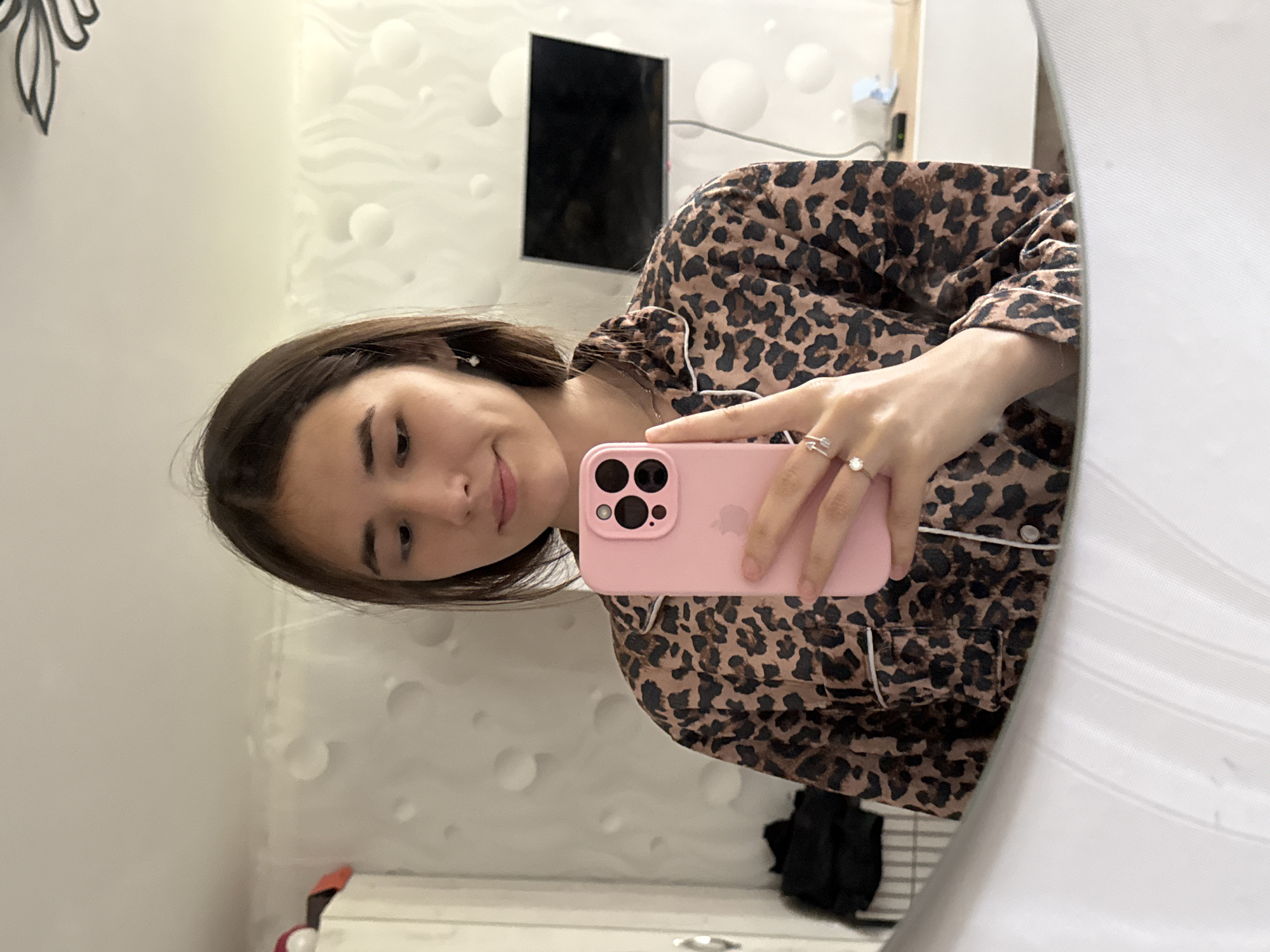 A young woman with dark brown hair is reflected in a mirror. She is wearing a leopard print pajama top with white trim. She is holding a pink smartphone in her right hand and has a ring on her left ring finger. Her eyes are looking down at the phone. In the background, there is a white textured wall, a television mounted on the wall, and a closet. The scene appears to be indoors, likely in a bedroom, in a residential setting in Chaikovsky, Russia.