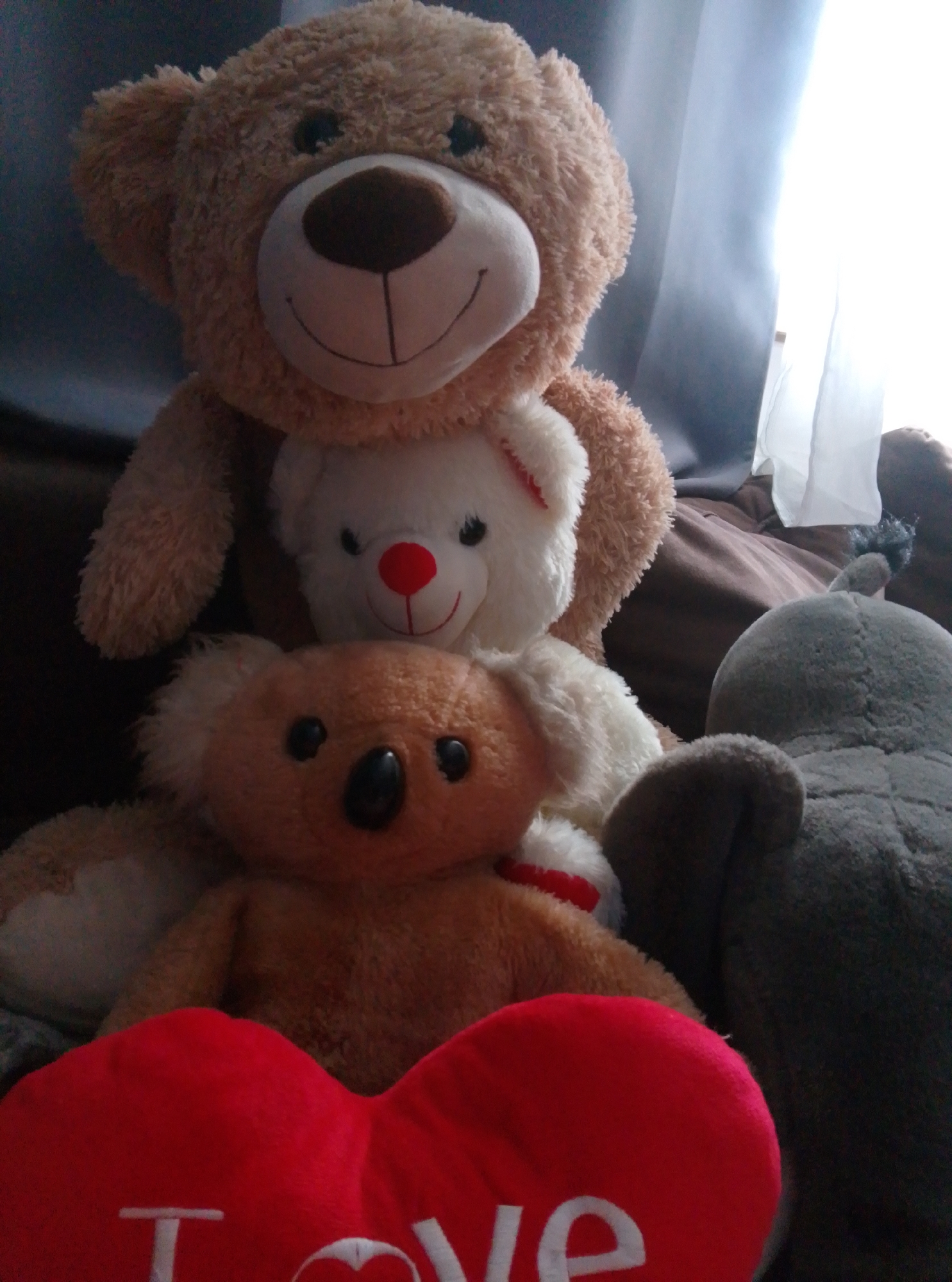 A group of stuffed animals is arranged on a brown sofa in front of a window with sheer white curtains. The largest stuffed animal is a light brown teddy bear, with a darker brown snout, eyes, and inner ears. It is positioned at the top of the arrangement. Below it is a smaller white teddy bear with a red nose and black eyes. In front of the white bear and slightly to the left is a brown koala bear with black eyes and a black nose. To the right of the koala bear and further back is a gray stuffed elephant. In the foreground and partially obscuring the lower stuffed animals is a large red heart-shaped pillow with the word "love" embroidered in white. The overall impression is that of a staged display of toys, possibly for a photograph. The image quality is moderate, with some blurriness and low light conditions. The specific location in the city of Wedel, Germany, is not discernible from the visual information provided.