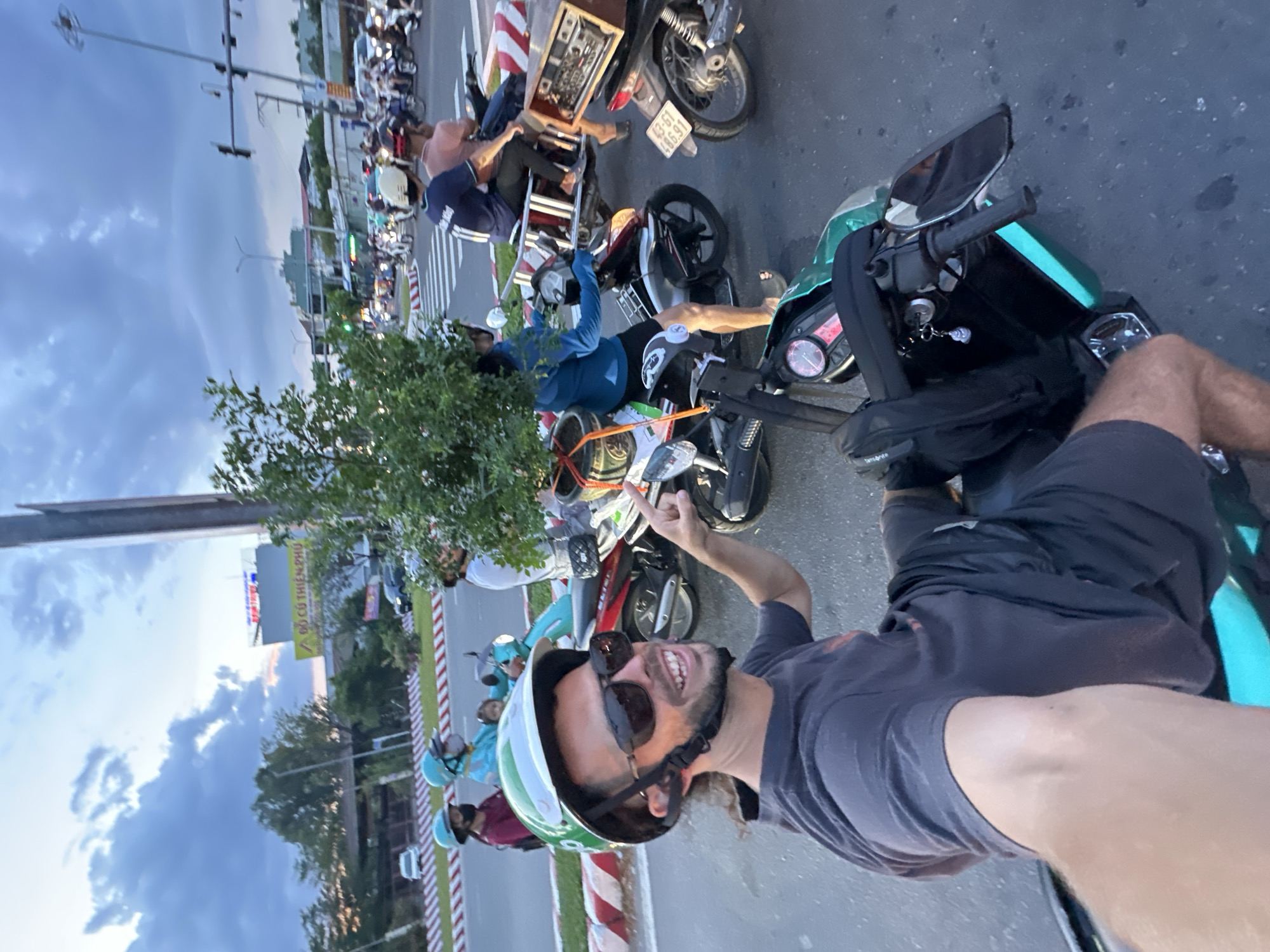 The image captures a selfie taken by a man with sunglasses and a green helmet on a motorcycle in Da Nang, Vietnam. He is smiling and pointing with his right hand. Behind him, other motorcycles and people are visible, suggesting a busy street scene. The environment includes traffic lights, street signs, and buildings in the background, indicating an urban setting. The sky is partly cloudy, suggesting it might be dusk or dawn. The overall impression is one of everyday life and movement in a Vietnamese city.