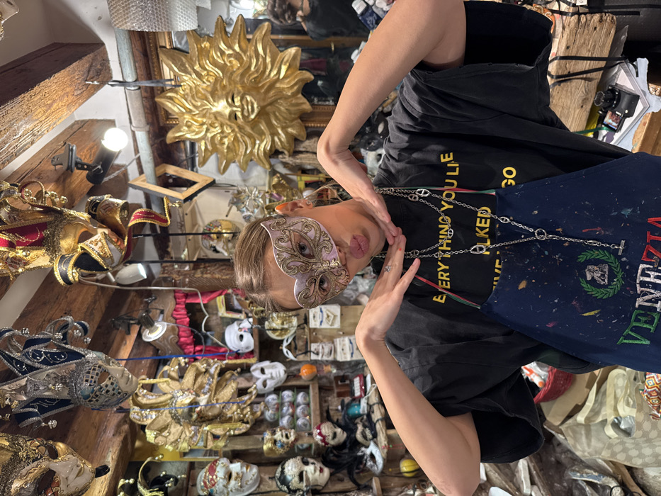 A young female stands centered in what appears to be a Venetian mask artisan workshop or specialized retail store in Venice, Italy. She wears a light pink and gold ornate masquerade mask that covers her upper face, an oversized black t-shirt with partially visible yellow text, and a paint-splattered dark blue apron featuring "VENEZIA" embroidered in green and red. Two long, silver-colored chain necklaces hang around her neck. Her hands are raised, with palms facing forward and thumbs touching her jawline, framing her face as her lips are pursed forward. The background is densely packed with a vast array of Venetian carnival masks, varying in style, color, and size, hanging from wooden beams and displayed on shelves. A large, golden sun-faced mask is prominently mounted on the wall to the right. The cluttered environment includes exposed wooden beams, various workshop materials, and a partial reflection of another individual's head in a dark surface on the far right.