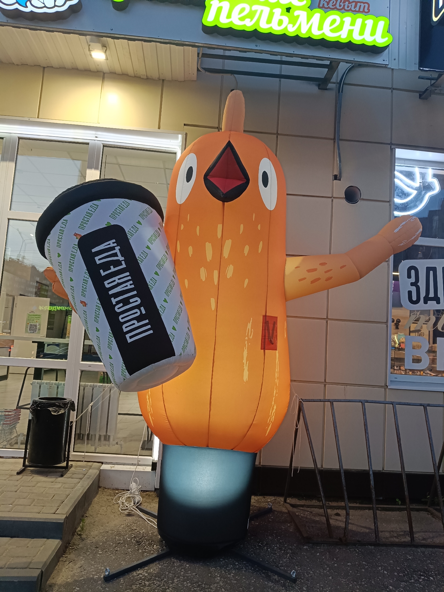 This is a medium, eye-level, full-shot photograph of an inflatable chicken mascot holding a large cup. The mascot is orange and has a cartoonish design with a large head, round eyes, and an open beak. It is standing on a black base that is lit from below. The cup it is holding is white with a black band and has repeating text in green that reads "ПРОСТАЯ ЕДА" (Prostaya Eda), which translates to "Simple Food." The mascot is positioned in front of a building with large glass windows and an awning above. A sign with bright green neon lettering is visible above the awning, partially obscured by the mascot. The sign appears to read "пельмени" (pelmeni), a type of Russian dumpling. To the left of the mascot, there is a black trash can and a shopping cart. The ground is paved with asphalt and concrete. The lighting suggests it is evening or early morning. The scene is outdoors, likely in front of a store or restaurant.