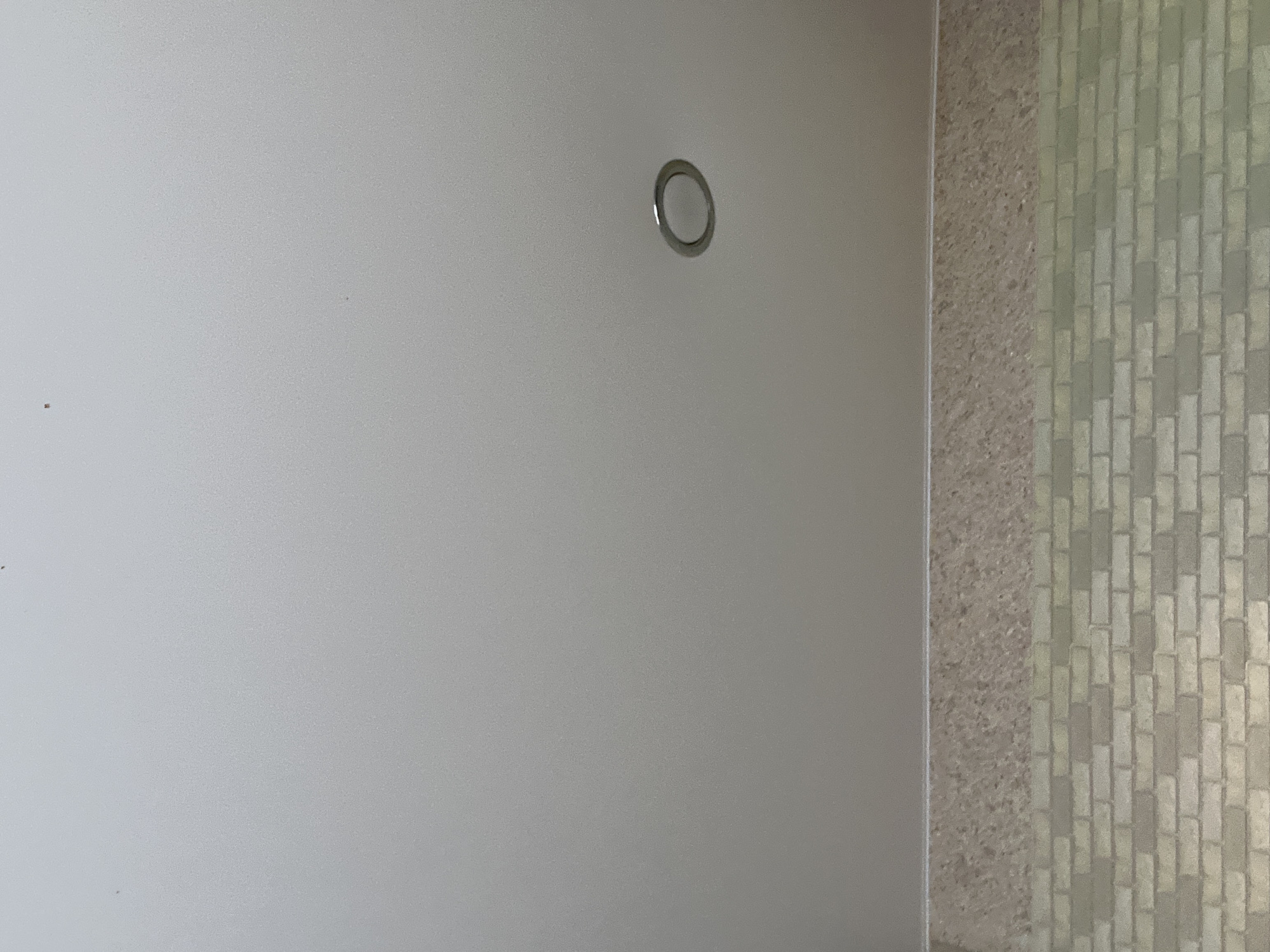 The image shows a section of a wall. The left side of the wall is a flat, light gray color. There is a small, circular metal fixture on this section of the wall. The right side of the wall features a textured surface composed of small, rectangular blocks arranged in a pattern resembling bricks. The exact location in Chaikovsky, Russia, cannot be determined from this image.