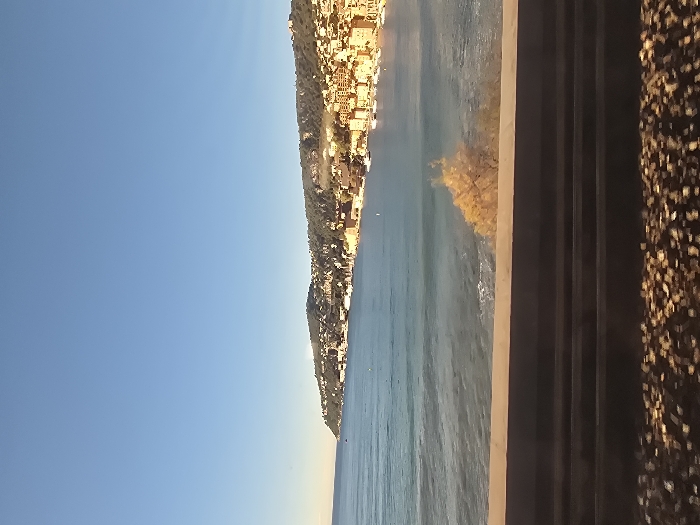 The image presents a panoramic coastal landscape, consistent with Villefranche-sur-Mer, France, captured from an elevated perspective on a bright, sunny day. A vast, clear blue sky dominates the upper and left portions of the frame, indicating excellent weather conditions.
Below the expansive sky and extending across the middle of the scene, a charming Mediterranean town is nestled into a verdant hillside. Its light-colored buildings are densely packed and cascade down towards the calm, blue-green waters of the sea. The sunlight brightly illuminates the town's facades and the surrounding hillside vegetation, suggesting a late morning or early afternoon setting.
In the immediate foreground, spanning the right side of the image, is a wide, dark grey structure. This appears to be a multi-lane road, highway, or a substantial promenade, featuring several distinct horizontal lines or sections running parallel to the coast. Adjacent to this structure, further to the right, is a textured, brownish-gold surface, possibly an embankment or shoulder covered in gravel or small stones, which glistens under the direct sunlight. Some patches of yellowish-brown foliage are visible along the coastline and dotting the hillside, adding to the natural beauty.
The sea exhibits gentle ripples and a soft shimmer, reflecting the overhead sunlight. There are no visible people, animals, or discernible text in the image. The clear skies, bright sunlight, and calm sea convey a serene, peaceful, and picturesque atmosphere.