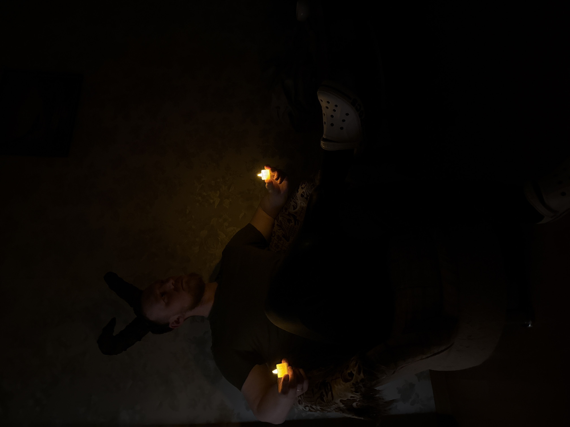 A person wearing horns holds two small, battery-operated candles in a dark room. The room appears to be a domestic setting. There are Crocs in the room and a sofa visible. There is no evidence in the image to identify the location as the city of Perm, Russia.