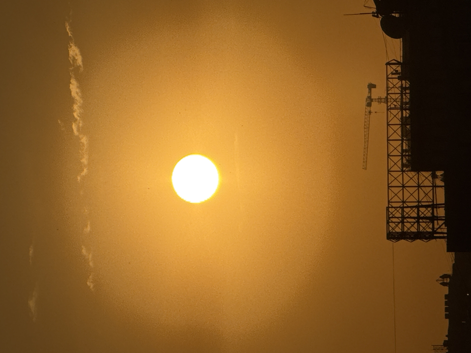 The image captures a bright, slightly hazy sun setting over a cityscape, casting an orange glow across the sky. In the foreground, silhouetted against the sky, are several structures. To the left, there's a dark, angular framework that appears to be part of a building or scaffolding. In the center, a construction crane is visible, suggesting ongoing development in the area. Further to the right, other dark, indistinct building shapes and a parabolic dish antenna are present. Two small, dark figures, possibly people, are standing on a structure to the left of the crane. The overall impression is one of urban development against a dramatic sunset.