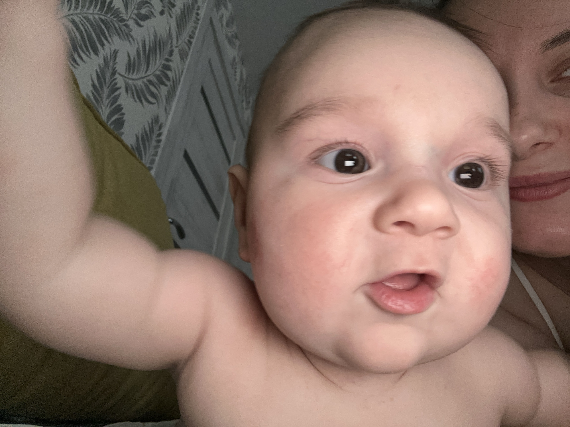 A baby with light skin is centered in the frame, their face filling the space. The baby has dark eyes, a slightly open mouth revealing the tip of their tongue, and chubby cheeks with some redness. The baby's left arm is raised and extends out of the frame on the left side.
A portion of an adult woman's face is visible in the upper right corner of the image. She also has light skin and her lips are visible.
The background includes a headboard with a gray and white leaf pattern. A yellow cushion is visible to the left of the headboard.
The scene appears to be indoors. The city of Chaikovsky, Russia is not identifiable.