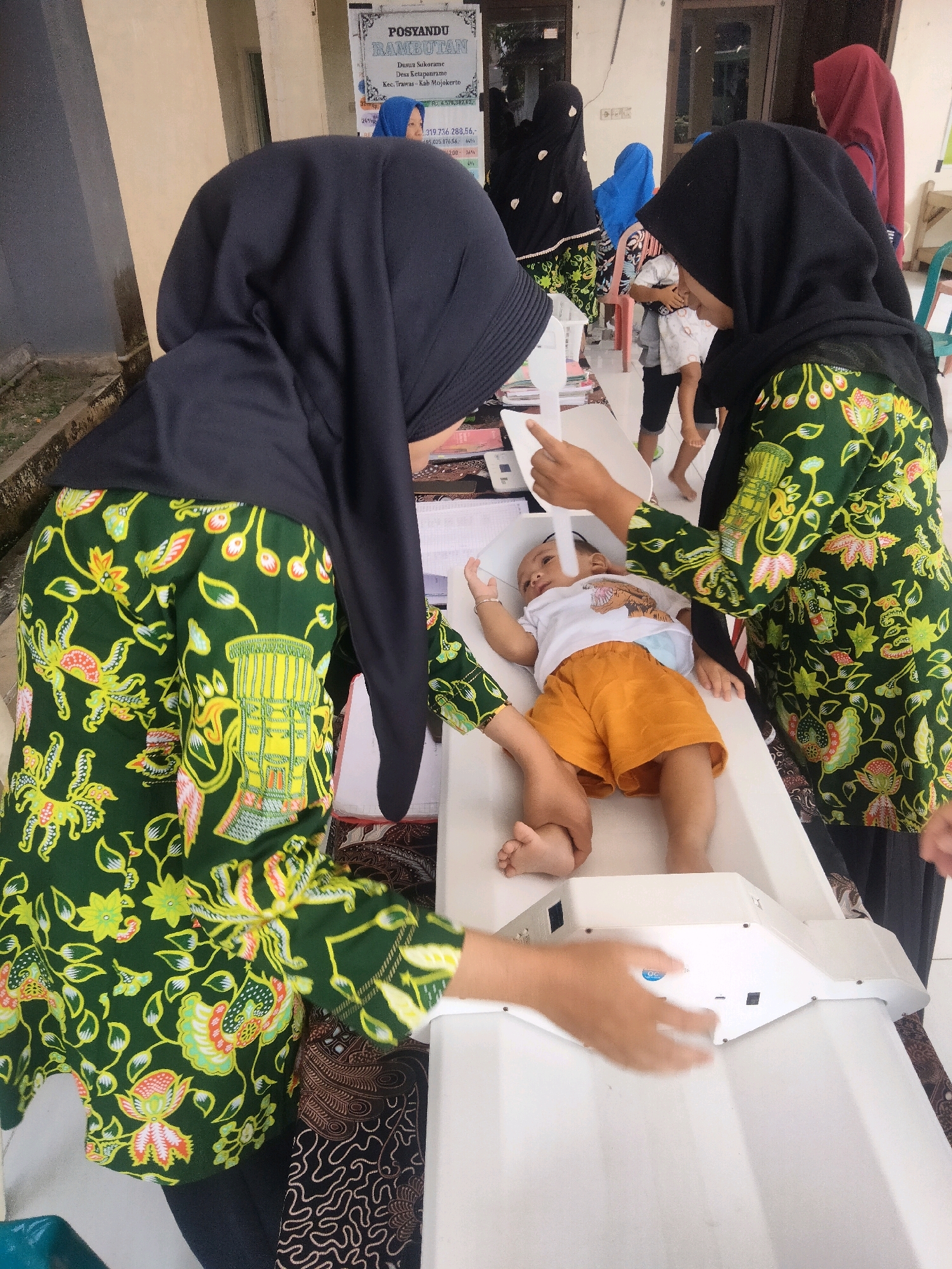 The image depicts a scene at a Posyandu (integrated health post) in Tretes, Indonesia. Two women, dressed in dark hijabs and vibrant green batik tops, are attending to a baby lying on a white scale. The woman on the left is partially obscured but appears to be adjusting the scale or holding the baby. The woman on the right is holding a white measuring device over the baby, who is wearing an orange outfit. Other individuals, including a child in a blue hijab, are visible in the background. A sign labeled "POSYANDU RAMBUTAN" is mounted on a wall behind them. The environment appears to be an indoor setting with a neutral-colored wall and some wooden elements.