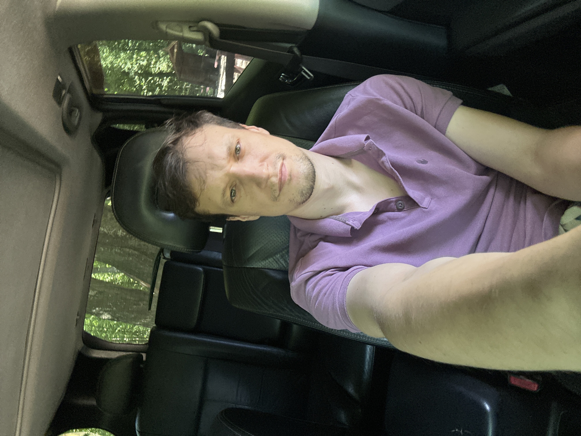 A man is sitting in the driver's seat of a vehicle, taking a selfie. The vehicle appears to be parked, possibly in a wooded area given the trees visible through the rear windows. The man is wearing a purple polo shirt. The interior of the vehicle is visible, with black leather seats and a sunroof. The overall lighting suggests it is daytime. There is no specific indication of the city of Shatava, Ukraine, or any other specific location beyond what can be inferred from the environment.