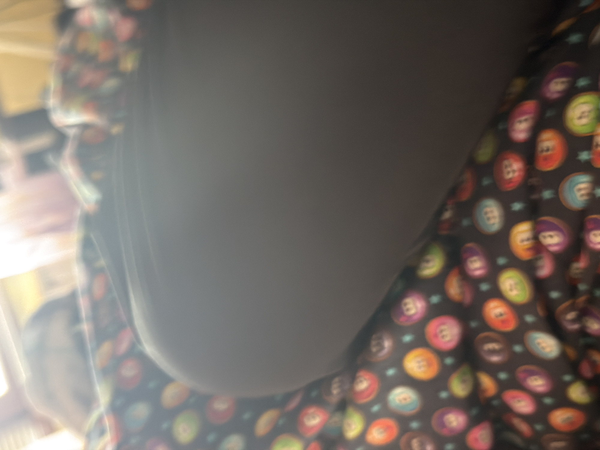 The image presents a blurry, close-up view, likely taken indoors in Iskar, Bulgaria. The central subject appears to be a person, with a large, plain dark grey area dominating the composition, possibly representing a torso, arm, or a piece of solid-colored clothing. This area is smooth and catches light, suggesting a fabric texture.
To the right and wrapping around the lower-left, a distinct patterned fabric is visible. This fabric is dark grey or black and features numerous colorful, circular designs. These circles strongly resemble billiard balls, each displaying a number (such as 8, 3, 13, 10, 15, 1, 9, 2, 7, 6, 4, 14, 12, 11, 5) in various hues like red, orange, green, blue, purple, and yellow. Small, light blue star-like shapes are interspersed between the circular patterns.
The lighting suggests an indoor setting, with a strong, possibly artificial, light source originating from the upper-left, causing a bright flare and highlights on the plain dark grey surface. The background on the far left is extremely out of focus, showing only indistinct light and shapes that could be furniture or parts of a room. No specific activity is discernible due to the static nature and blurriness of the photograph. The perspective is unusual, possibly a spontaneous shot from an atypical angle. No specific location cues for Iskar, Bulgaria are discernible within the image itself.