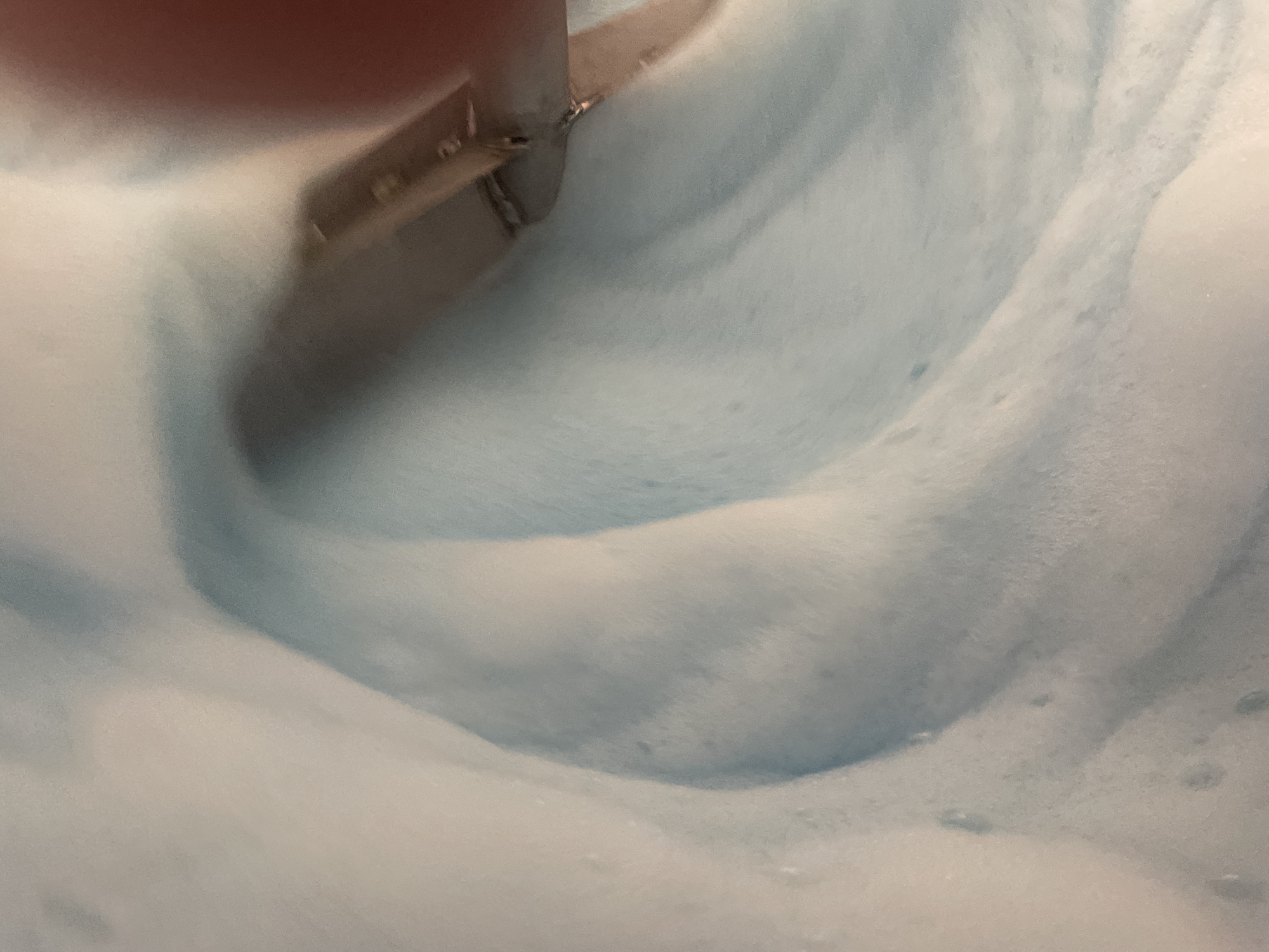 A close-up, top-down shot shows a light blue and white foamy substance, possibly suds or lather. The texture is soft and undulating, with a subtle swirling pattern. A metallic object, appearing to be part of a tool or utensil, is partially submerged in the foam on the right side of the frame. A blurry, reddish-pink element, likely a finger or hand, is also visible at the top right corner, suggesting human interaction with the substance. The lighting is diffuse, creating soft shadows and highlights on the foam. There are no discernible landmarks or environmental features that would indicate a specific location within the city of Терволово, Russia, or any other location.
