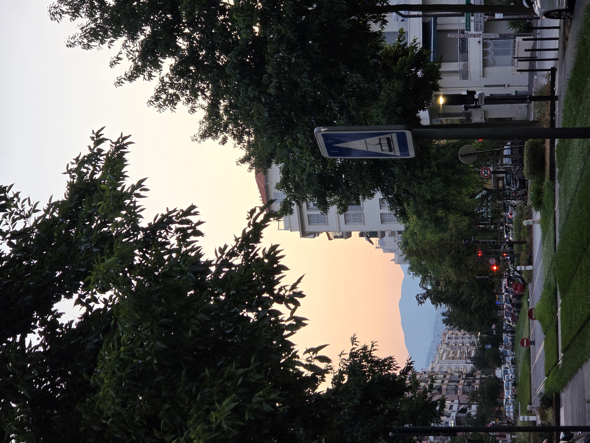 This is a vertical, eye-level, medium shot captured at dusk on a city street in Nice, France. In the foreground, dark green trees frame the left and right sides of the image, with their leaves silhouetted against a pale orange and yellow sky. A light pole with a blue and white sign depicting a tram is positioned in the mid-right foreground. The sign is partially obscured by the trees. Further down the street, a traffic light with red lights illuminated is visible. Buildings with balconies and windows line the street on the left, extending into the distance. On the right, more trees and buildings are present, including a white building with balconies. Cars are parked and driving on the street, and a light pole with a yellow light is visible between the trees on the right. In the background, a range of light blue mountains can be seen against the horizon. The street features tram tracks and green grass.