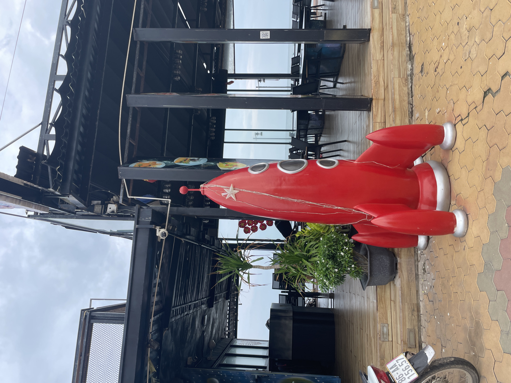 The scene is an outdoor setting in Phu Thuy Ward, Vietnam. A red, retro-style rocket statue adorned with string lights stands prominently on a patterned brick surface. The rocket appears to be part of the decor for a restaurant or cafe, suggested by the outdoor tables and chairs visible in the background, along with the steel beam framework of the structure. Potted plants provide some greenery. Part of a parked motorbike is visible in the lower right corner of the image. The sky is overcast.