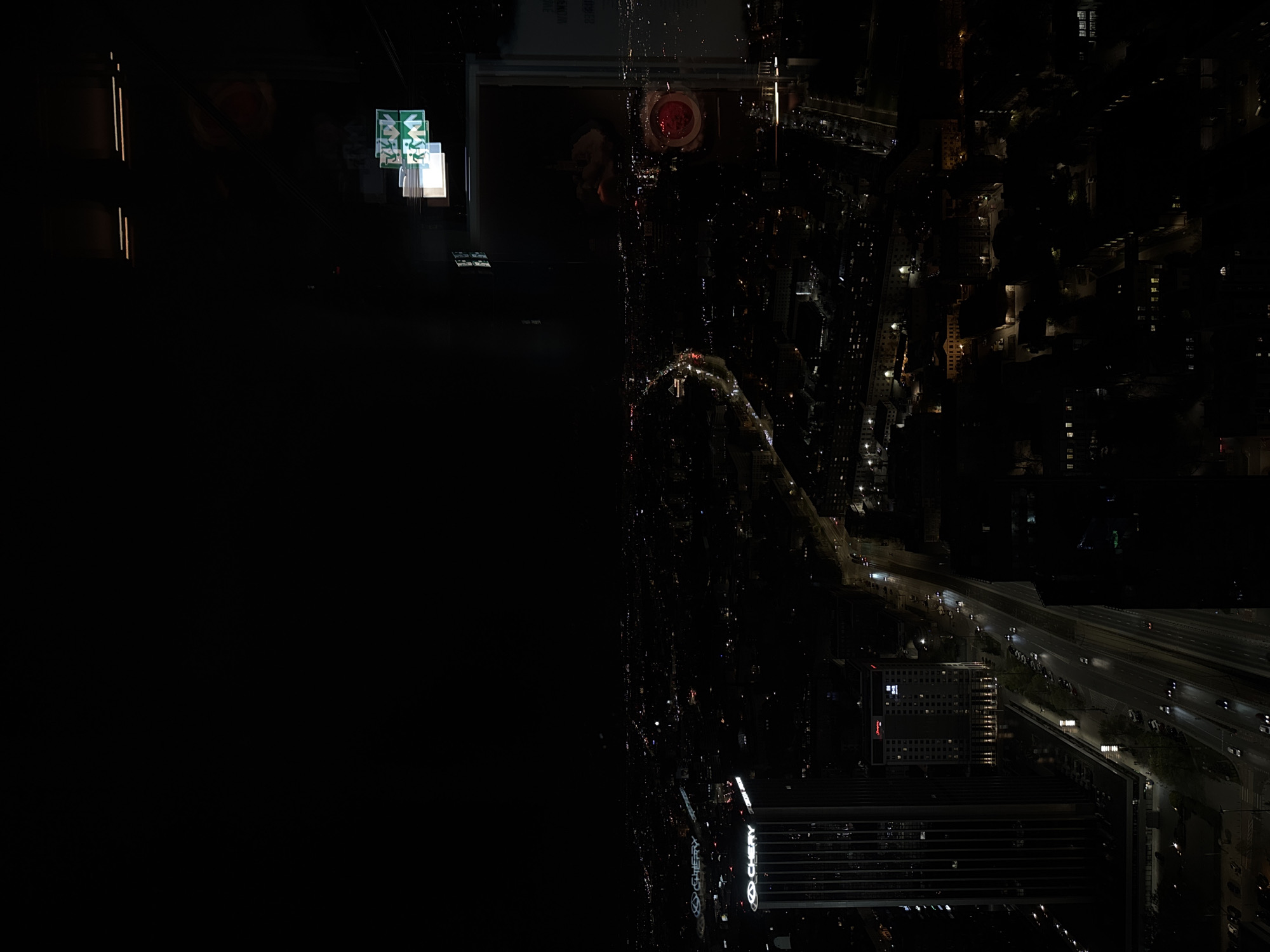 An aerial view of Warsaw, Poland, at night. The left side of the image is dark and obscured. On the right side, city lights illuminate the buildings and streets below. A highway runs along the bottom edge of the frame. The image is rotated 180 degrees. A Chevy sign is visible.