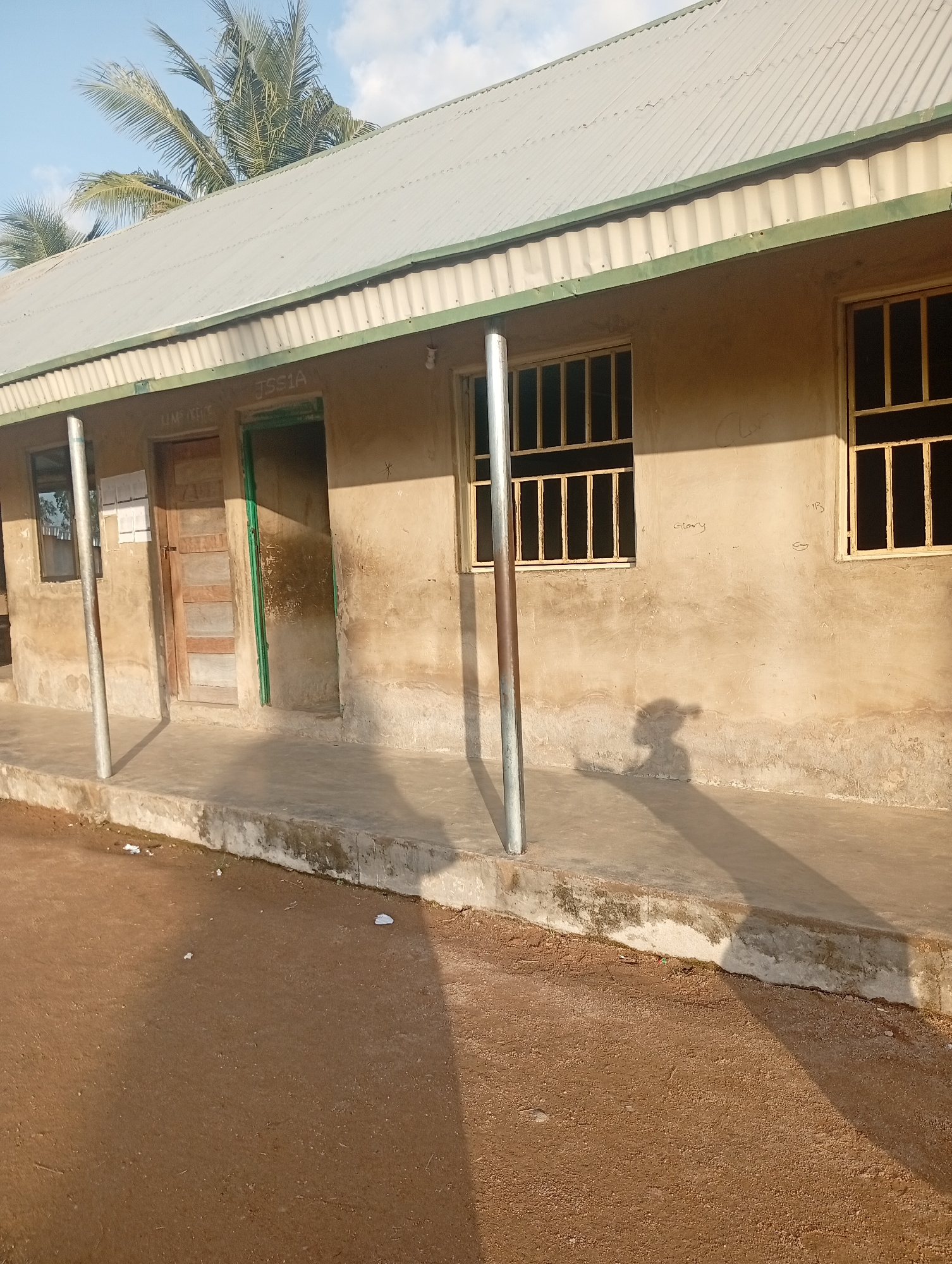 The image depicts the exterior of a modest, single-story building, likely a school, in Jalingo, Nigeria. The building has a corrugated metal roof with a green trim along the eaves. The walls are made of weathered, light-colored stucco, showing signs of age and possibly some water damage near the base.
There are several windows visible, all fitted with yellow metal bars for security. To the left, a window is partially obscured by a tree's reflection. Next to it is a wooden door painted in a light brown hue, with vertical planks. To the right of the door is an open doorway, framed in green, leading into the interior of the building.
A metal pole stands vertically in front of the building, casting a long shadow on the concrete walkway that runs along the front. The ground in front of the walkway is covered in reddish-brown dirt. The shadow of a person, possibly a child, is clearly visible on the walkway, suggesting they are standing somewhere out of frame.
The sky above is blue with some scattered white clouds, indicating fair weather. A palm tree is visible in the upper left corner of the image, its fronds reaching over the roof, hinting at a tropical or subtropical climate.
Visible text includes "JSS1A" written in white paint above the doorway. There are also faint markings and what appears to be graffiti on the wall, including the word "glory." A few pieces of white paper are scattered on the ground near the walkway. The overall impression is of a functional but somewhat aged educational facility in a warm climate. The time of day appears to be late afternoon, given the length and angle of the shadows.