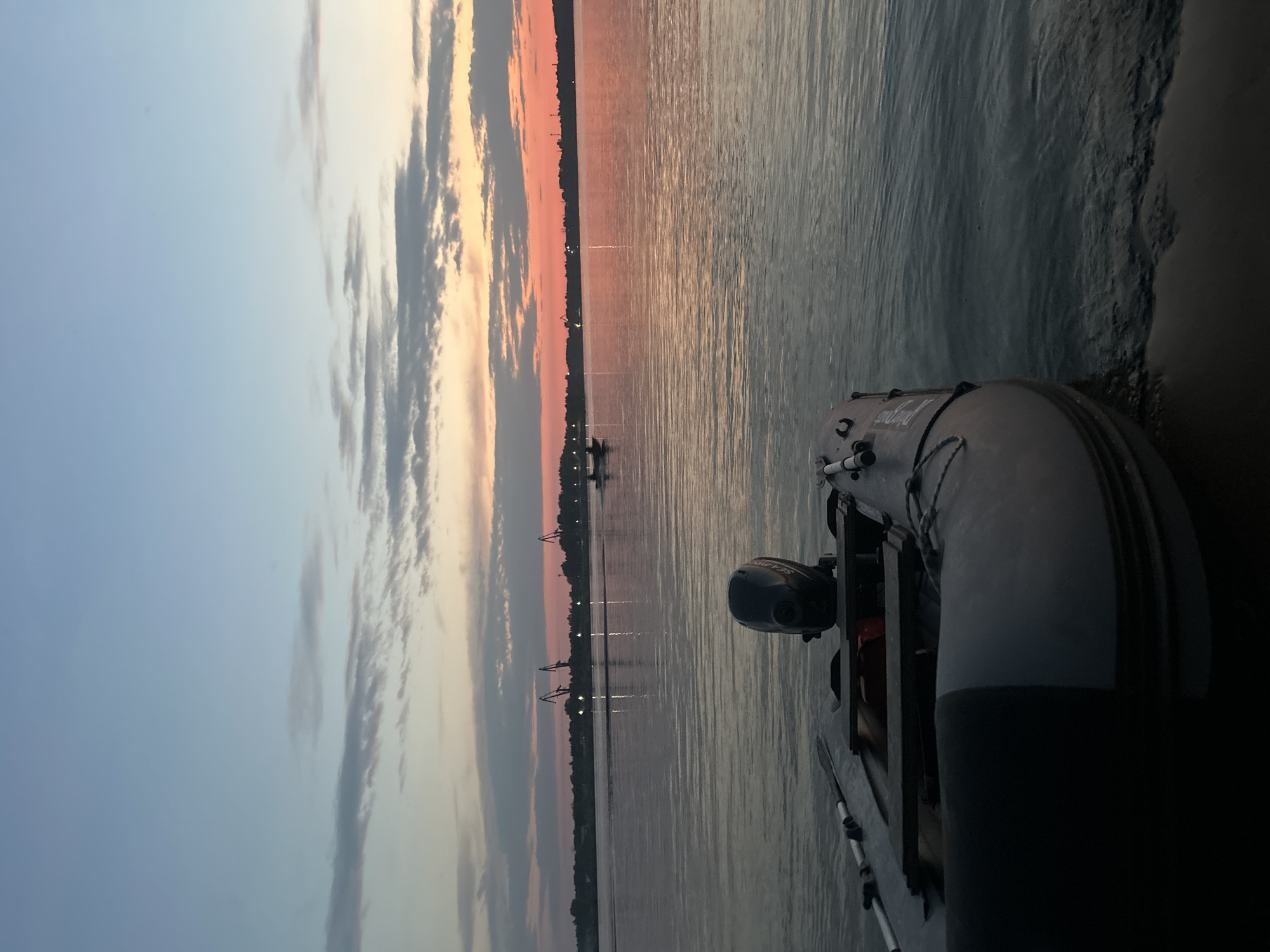 A twilight view captures an inflatable boat with an outboard motor moored at the edge of a calm body of water. In the distance, the silhouette of a city skyline is visible, featuring industrial cranes and lights reflecting on the water. The sky displays a gradient of colors from light blue at the top to vibrant orange and pink hues near the horizon, indicating either sunrise or sunset. Two smaller boats are also present on the water further away, and the overall atmosphere is serene. The scene is situated in the city of Chaikovsky, Russia, likely along the Kama River.