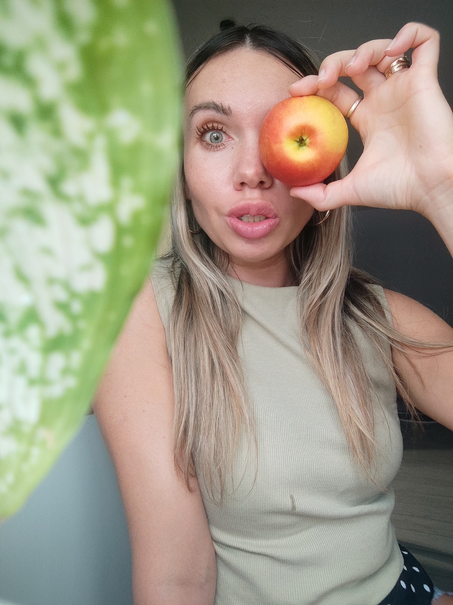 A young adult woman with fair skin, long blonde hair, and light-colored eyes is posing for a selfie. She is holding a small, red and yellow apple over her right eye. She has several rings on her right hand. She is wearing a light green ribbed tank top and is partially visible against a neutral gray-toned background. To the left, a large green plant leaf fills a portion of the frame. The lower part of the woman's attire reveals a polka dot pattern, suggesting shorts or a skirt.
No information is available to determine if the scene is in Votkinsk, Russia.
