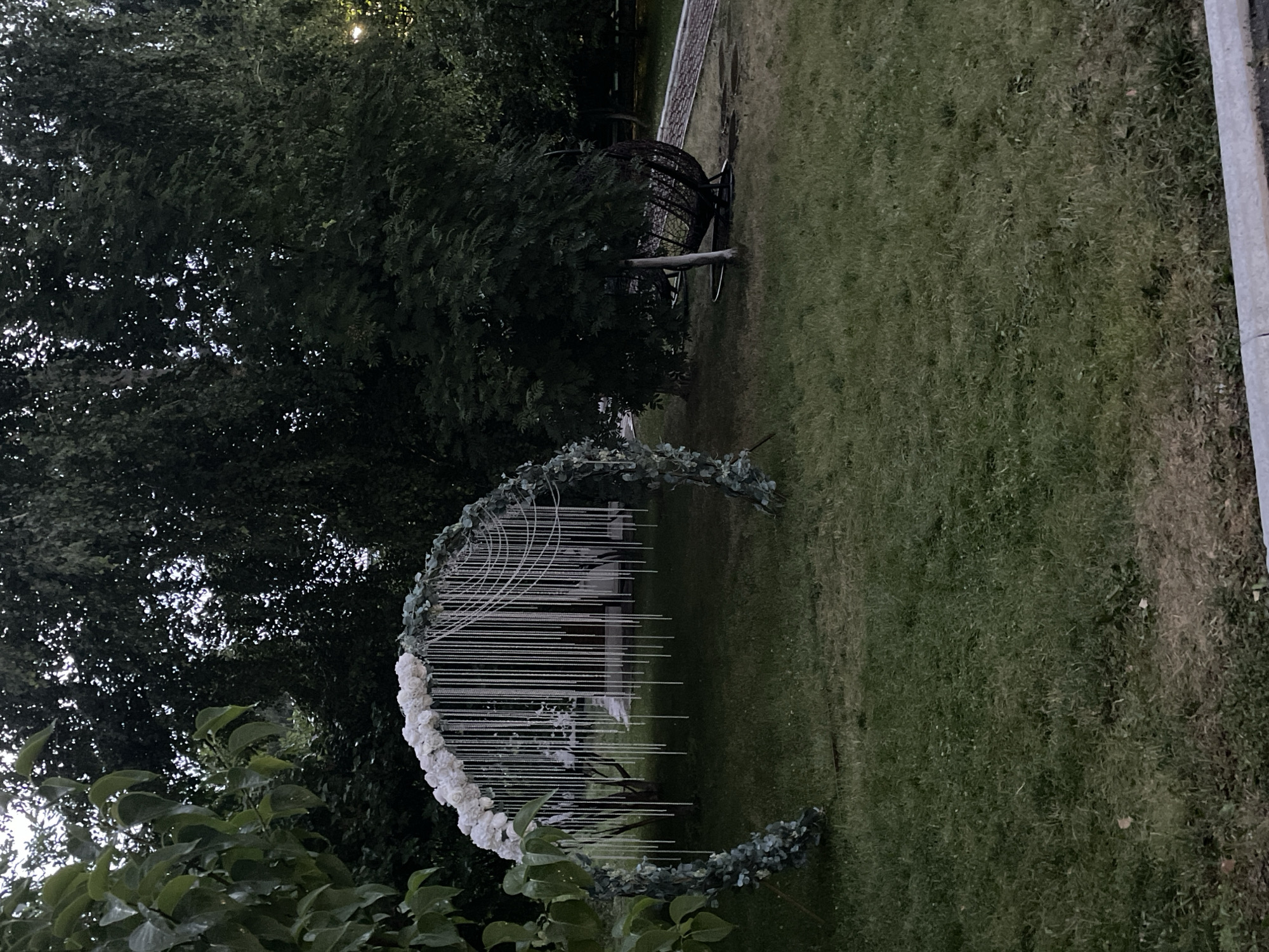 The image shows an outdoor scene, likely a garden or park, with lush green grass and dense trees. A circular wedding arch, adorned with white flowers and greenery, stands prominently in the foreground. The arch is made of a white, metallic structure with thin, hanging strands that create a delicate, airy effect. In the background, a large, dark, spherical object, possibly a decorative garden feature or seating area, is visible amongst the trees. The lighting suggests it is either early morning or late evening, with soft light filtering through the leaves. The overall atmosphere appears serene and tranquil, hinting at a peaceful setting, possibly for an outdoor event like a wedding or a gathering. There is no visible text in the image.