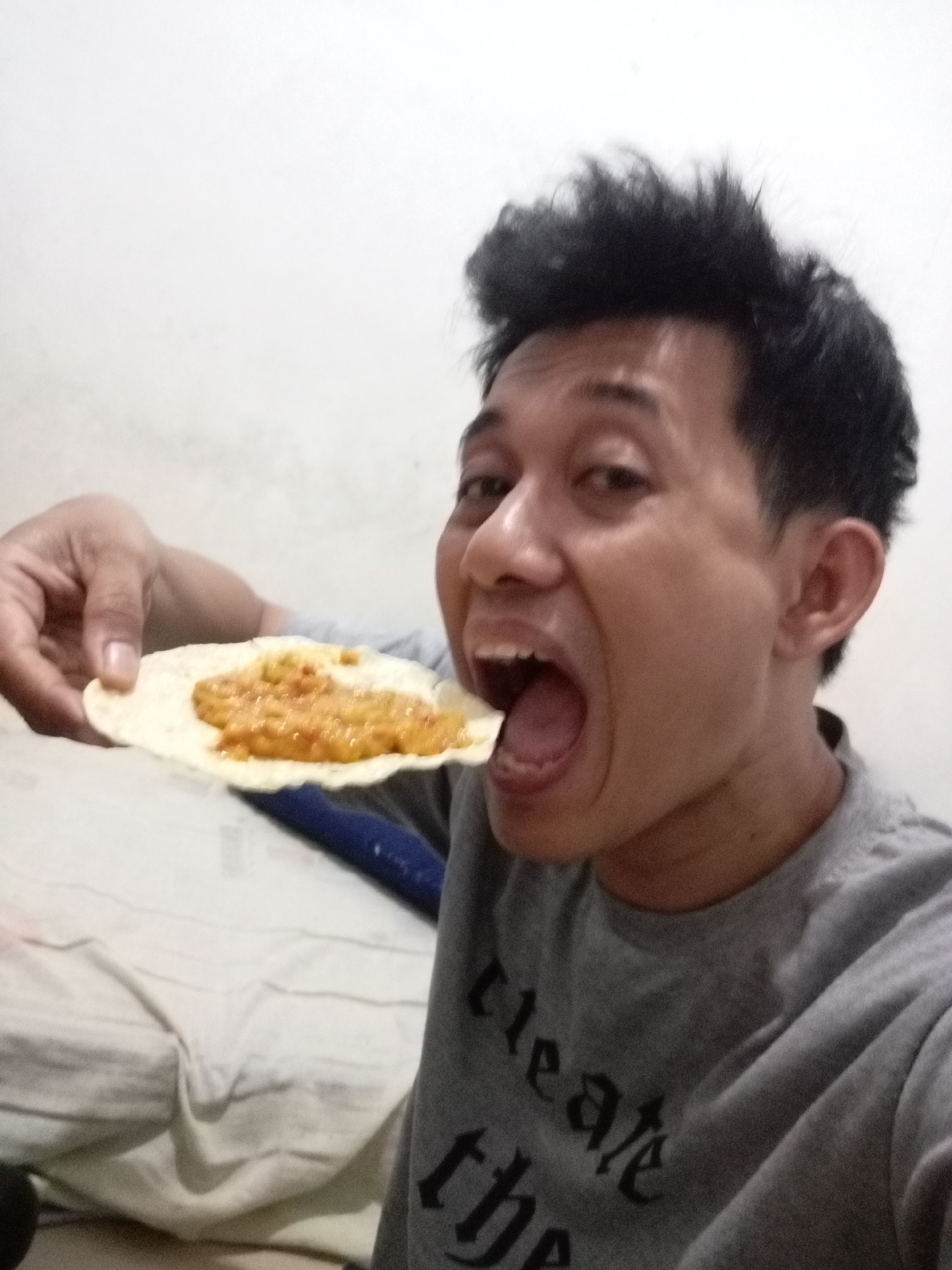A young Asian man is shown in a close-up, selfie-style shot as he prepares to eat a piece of flatbread topped with a yellow-orange condiment. His mouth is wide open, and the flatbread is being brought towards it by his left hand. The man has dark, spiky hair and is wearing a gray t-shirt with text that is partially obscured by folds in the fabric. The background consists of a lightly textured white wall and a portion of a light-colored surface that appears to be a bed with rumpled sheets. There is a dark blue object visible behind the man's shoulder. The lighting suggests an indoor setting. This scene likely takes place in Batam, Indonesia, given the available information about the image's context.
