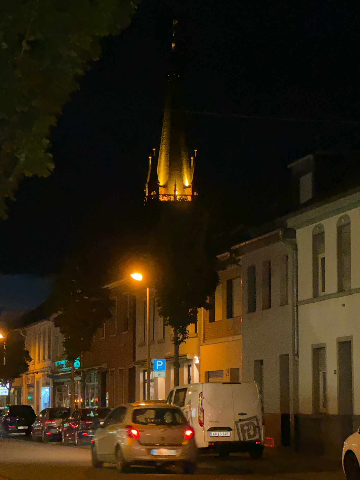 The image shows a street scene at night in Krefeld, Germany. In the background, a tall church spire is illuminated with warm light. In the foreground, cars are parked and driving along the street. Buildings line the street on the right side, with lights on inside some of them. A street lamp is visible on the left side of the image, also illuminated. The overall impression is of a quiet, urban street at night.