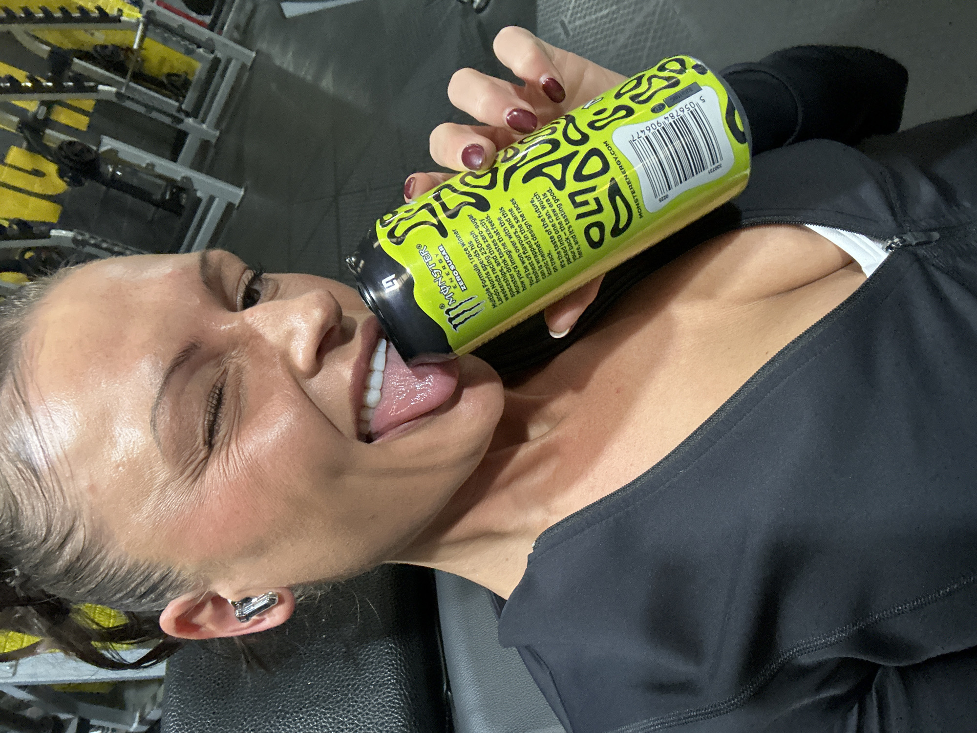 The image shows a woman lying on a bench in a gym. She is holding a yellow and black Monster Zero Sugar energy drink can to her mouth and sticking her tongue out to lick the top of the can. The can has a barcode and other text on it. She is wearing a black athletic top and has wireless earbuds in her left ear. Her fingernails are painted dark red. In the background, there is a weight rack with yellow weights. The text on the can includes the words "Multiple Formula 1 race winner Lando Norris" and "MONSTERENERGY.COM." No location is seen in the image.