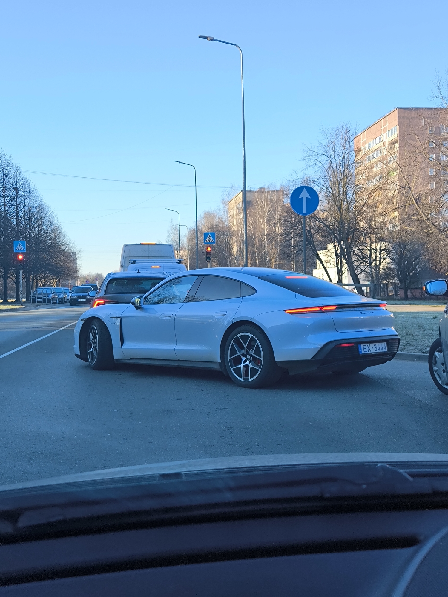 The image depicts a light blue Porsche Taycan, bearing the license plate "EX-3444", stopped in traffic on a city street. It is a daytime shot with a clear, blue sky. The traffic light visible in the background is red, indicating the vehicles are halted. Streetlights are present. There are other vehicles, including a white van, in the line of traffic. A pedestrian crossing sign is also visible. The image was taken from inside another car, looking through the windshield. The location is likely Riga, Latvia given that the license plate is a Latvian license plate. Buildings and bare trees line the street in the background. A blue, circular "one way" road sign is also visible.