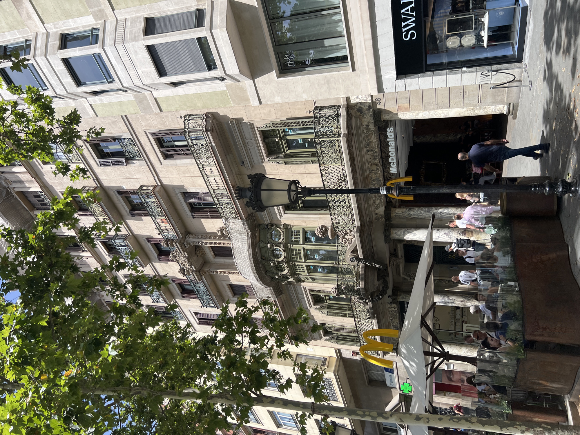 This media file depicts a street scene in Barcelona, Spain, on a bright, sunny day. The perspective is looking upwards at the facades of buildings, with a large tree in the foreground casting shadows. The architecture appears to be a mix of ornate historical styles and more modern elements.
The main subjects include several people, some walking on the sidewalk and others seated at outdoor cafe tables. The cafe area features a large white umbrella providing shade and a glass enclosure. Visible businesses include a McDonald's with its distinctive yellow arches and a Swarovski store with "SWAR" clearly visible on its signage.
The time of day is indicated by the strong shadows and bright sunlight, suggesting midday or early afternoon. The weather is clearly fair. The McDonald's and the presence of outdoor seating suggest a commercial area, likely a busy street. Some people are engaged in conversation or enjoying meals, indicating a relaxed, everyday atmosphere. A street lamp with an ornate design stands near the McDonald's. The overall mood is one of urban life on a pleasant day.