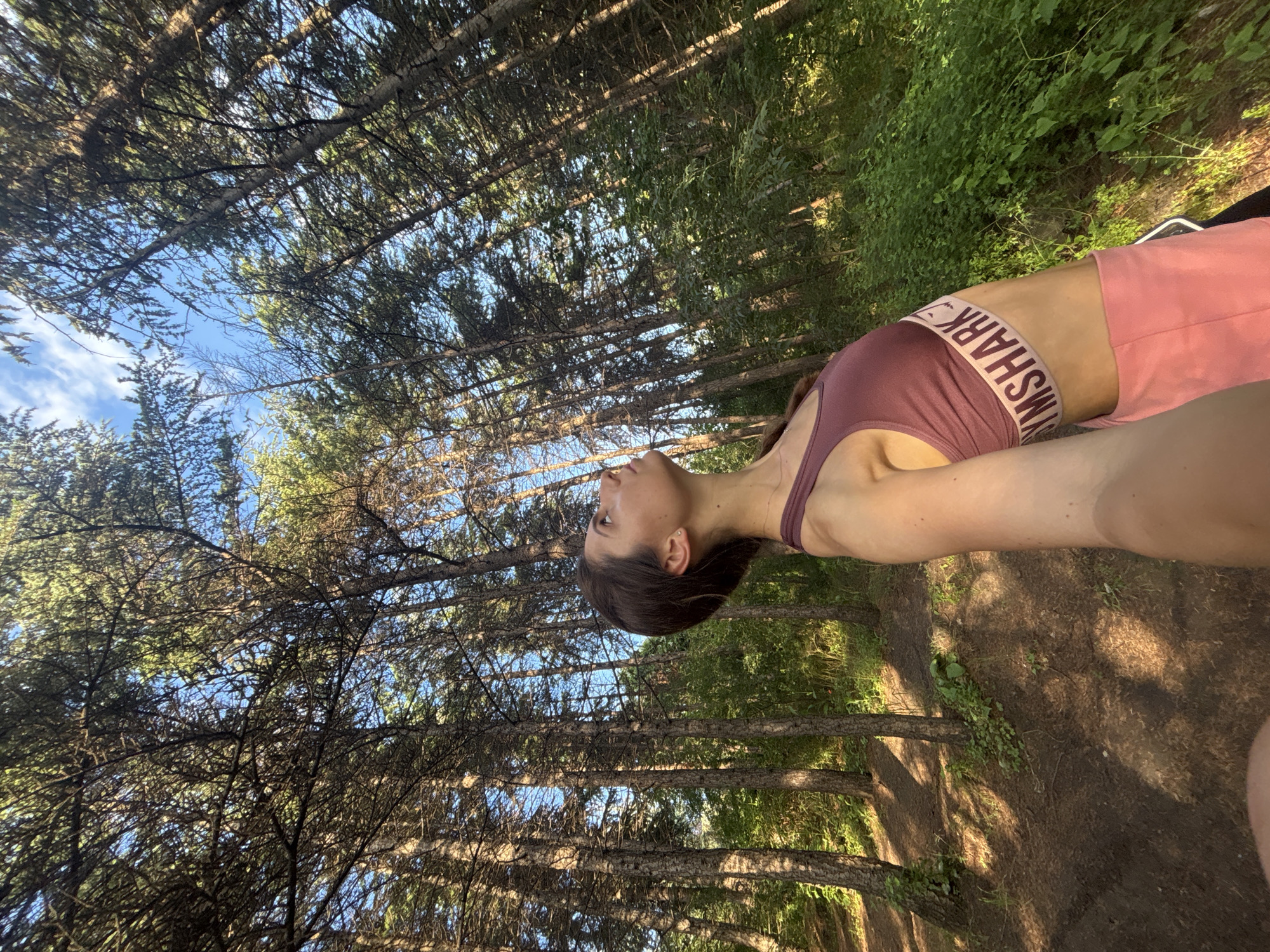 A woman is standing in a forest, looking up. She is wearing a maroon sports bra with a light-colored band that says "GYMSHARK" and pink shorts. Her right arm is extended, holding what is presumed to be a camera or phone to take a selfie. The trees are tall and slender, with a dense canopy of green leaves. Sunlight filters through the trees, casting dappled shadows on the forest floor. The ground is covered in dirt and some sparse green vegetation. The sky is blue with a few scattered white clouds. The overall impression is of a natural, outdoor setting. This image was taken in the city of Varna, Bulgaria.