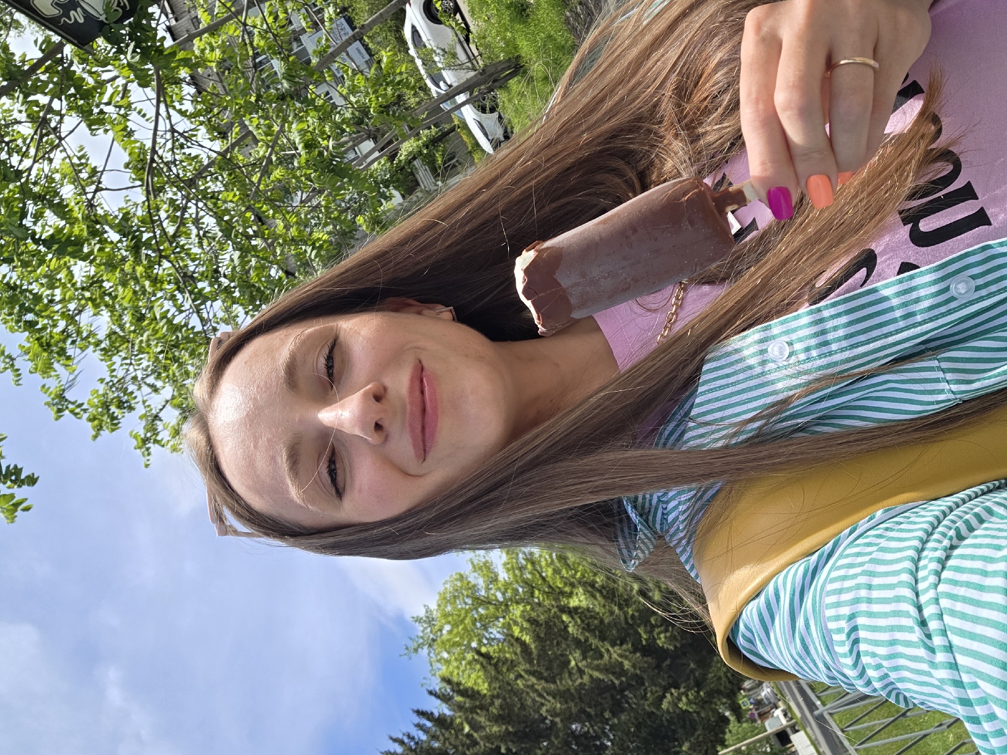 A woman is shown from a low-angle perspective, holding a partially eaten chocolate-covered ice cream bar in her right hand. She has long, light brown hair and is smiling slightly. She is wearing a pink t-shirt with writing on it and a green and white striped button-up shirt layered over the t-shirt, along with a yellow vest. Her fingernails on her right hand are painted different colors: pink and orange. She is wearing a ring on her right ring finger.
In the background, there are green trees and a blue sky. The location appears to be outdoors, possibly in a park-like setting. There are also vehicles visible in the background. The city of Birobidzhan, Russia cannot be conclusively identified from the visual information in this image.
